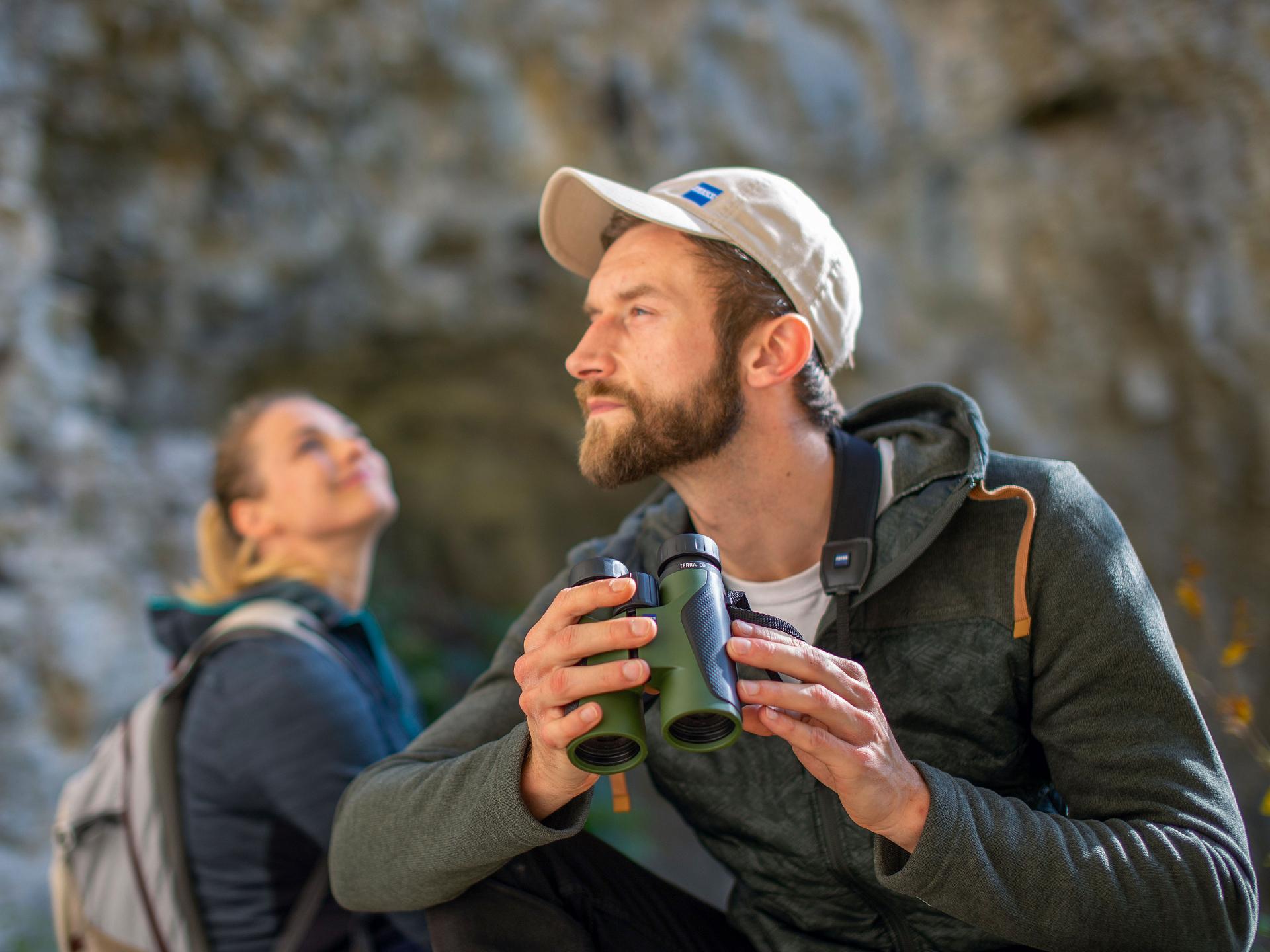 A man holding ZEISS binoculars sits outdoors next to a woman, both dressed for hiking in a rocky natural setting.