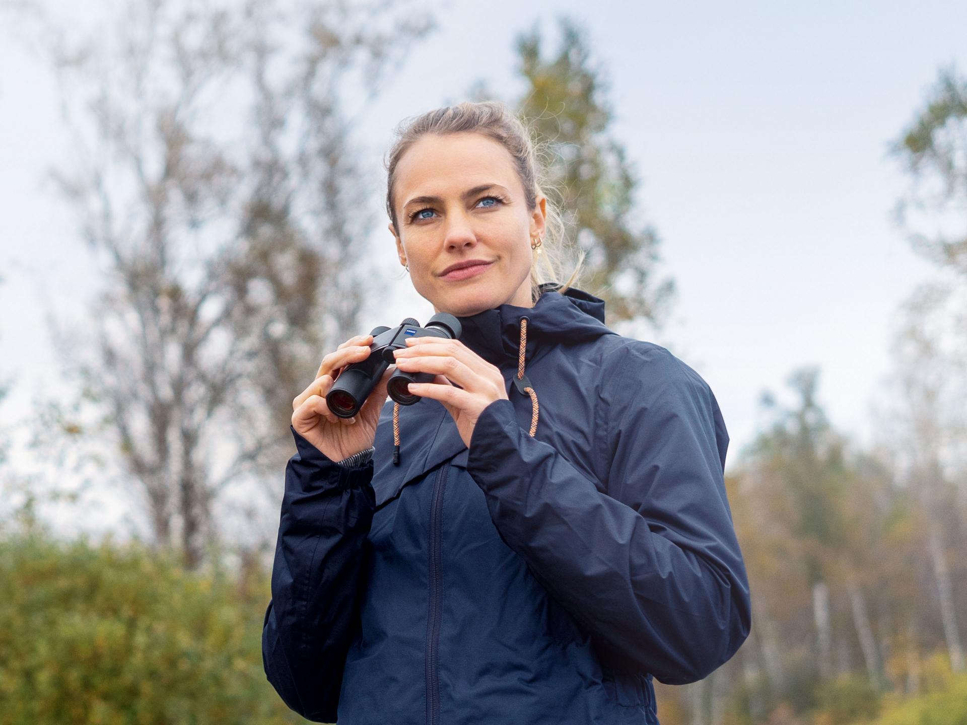 Woman wearing a navy outdoor jacket holding ZEISS binoculars in a forested area during daytime.
