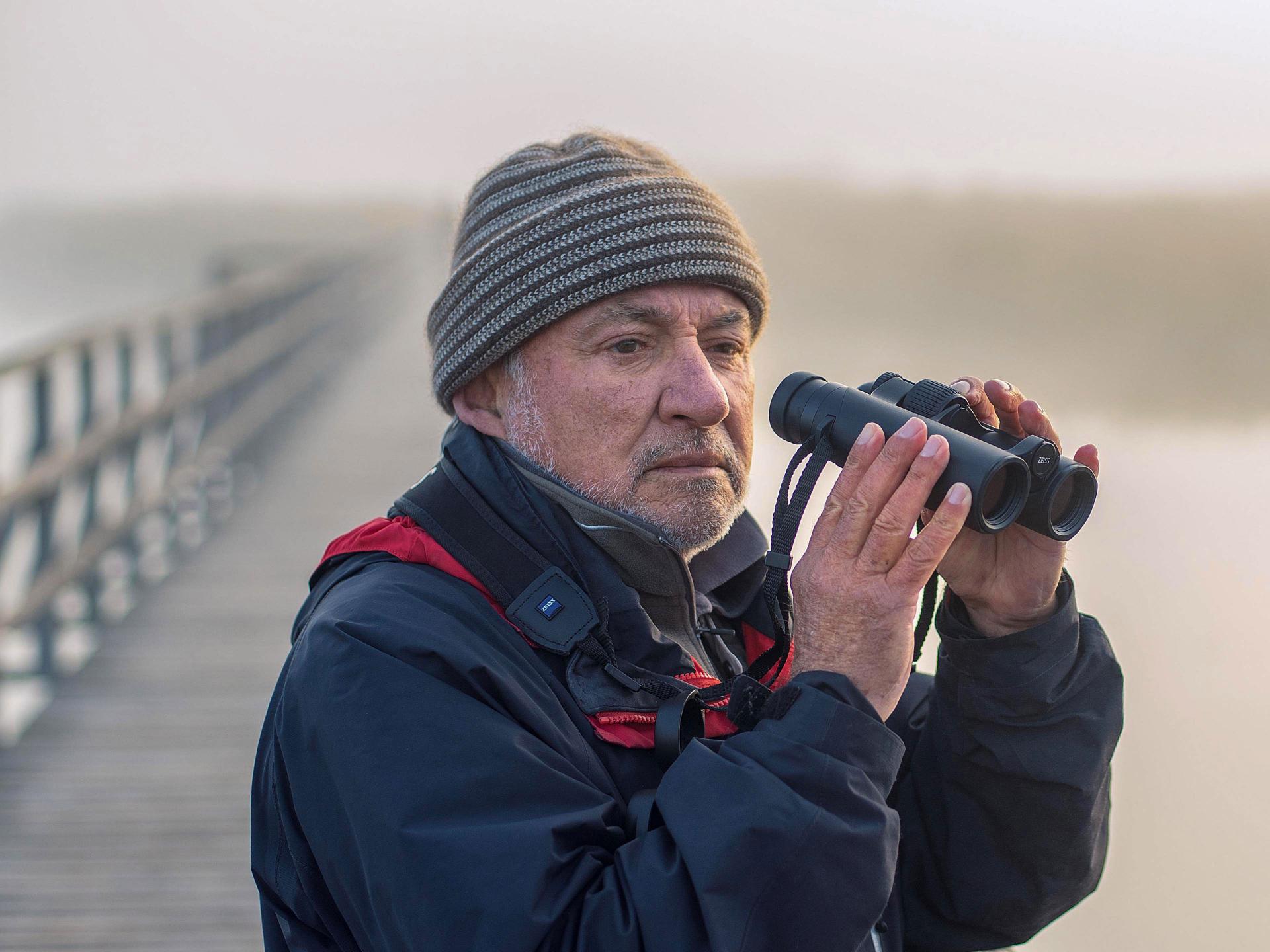 An older man wearing outdoor gear is holding a pair of ZEISS binoculars while standing on a foggy boardwalk.