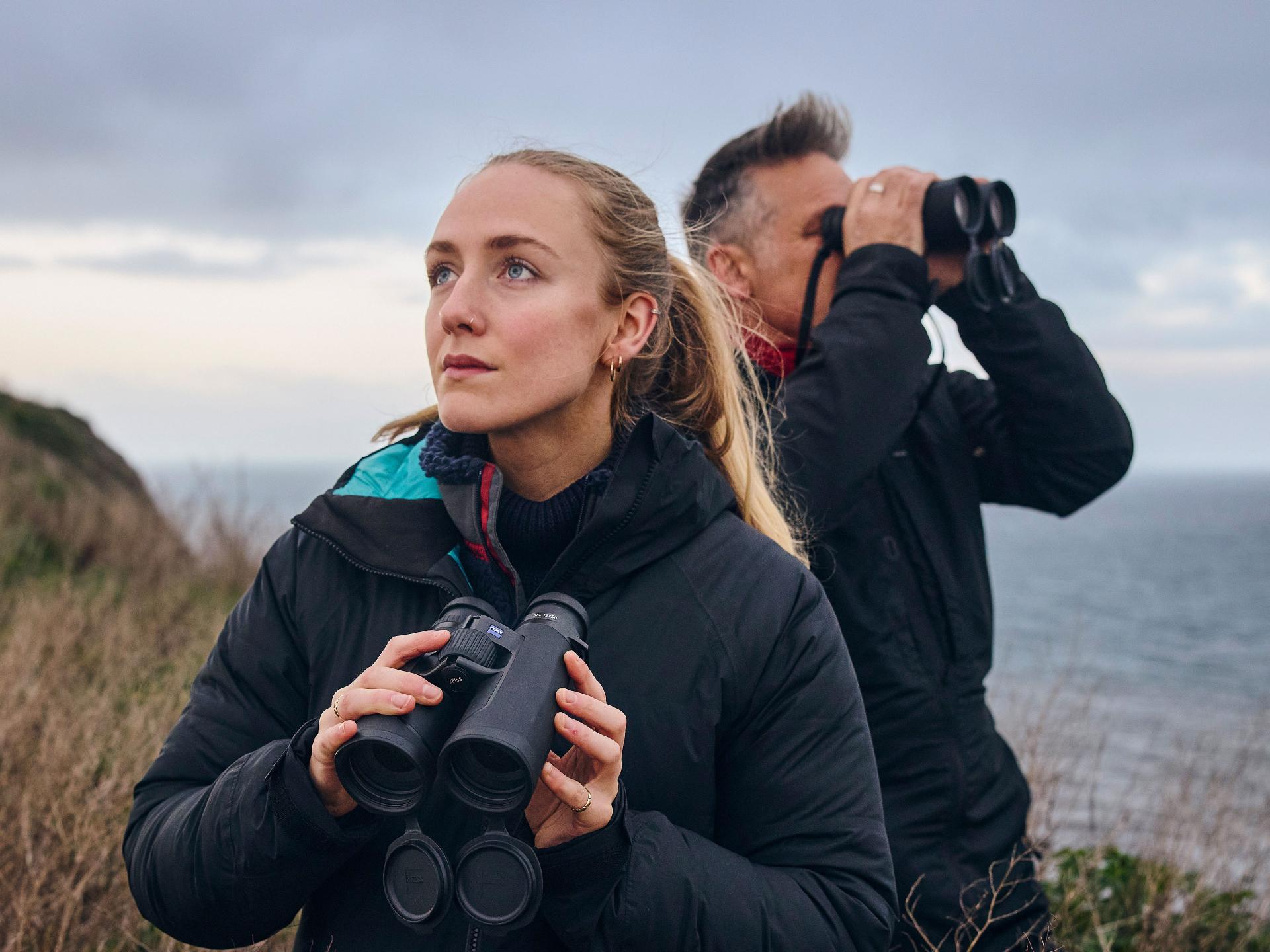 Two people stand on a coastal cliffside using ZEISS binoculars to observe the landscape.