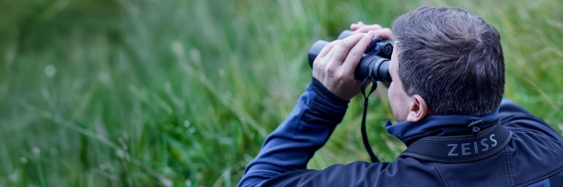 A person wearing a ZEISS carrying strap is observing nature through binoculars in a grassy outdoor setting.