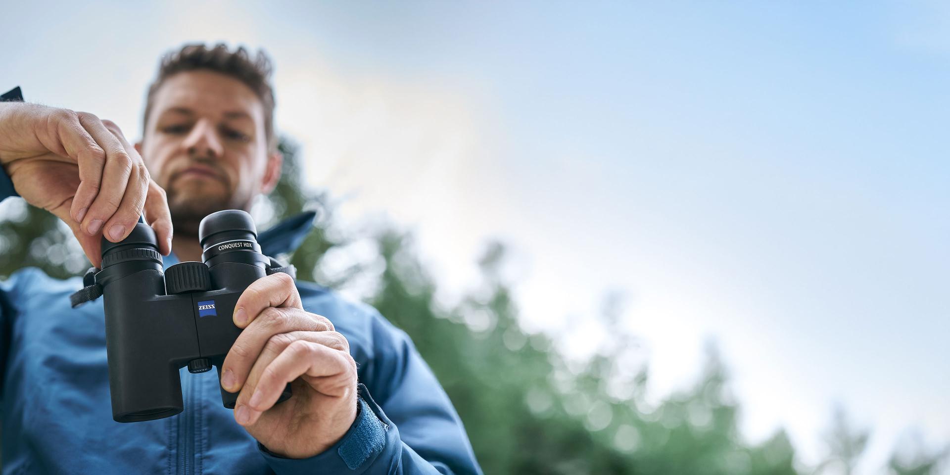 Man in a blue jacket adjusting a pair of ZEISS binoculars outdoors with trees and sky in the background.