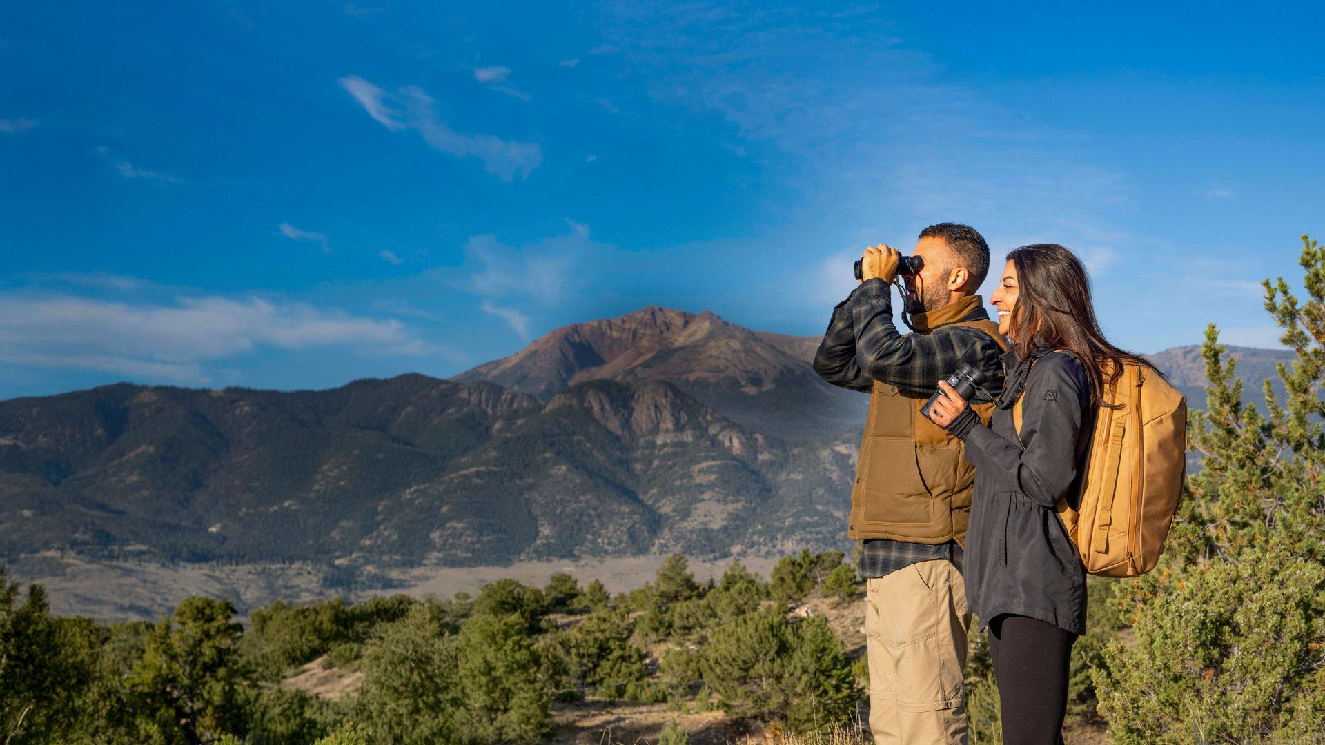 Two hikers stand in a scenic mountain landscape, with one using ZEISS binoculars to observe the distant view.