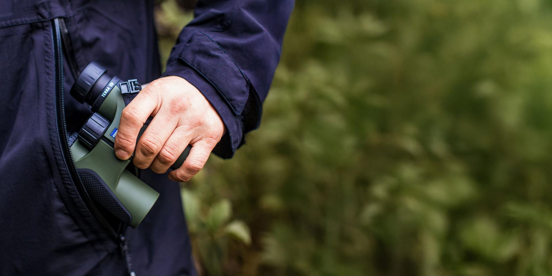 A person in a dark jacket is holding green ZEISS binoculars against a blurred natural outdoor background.