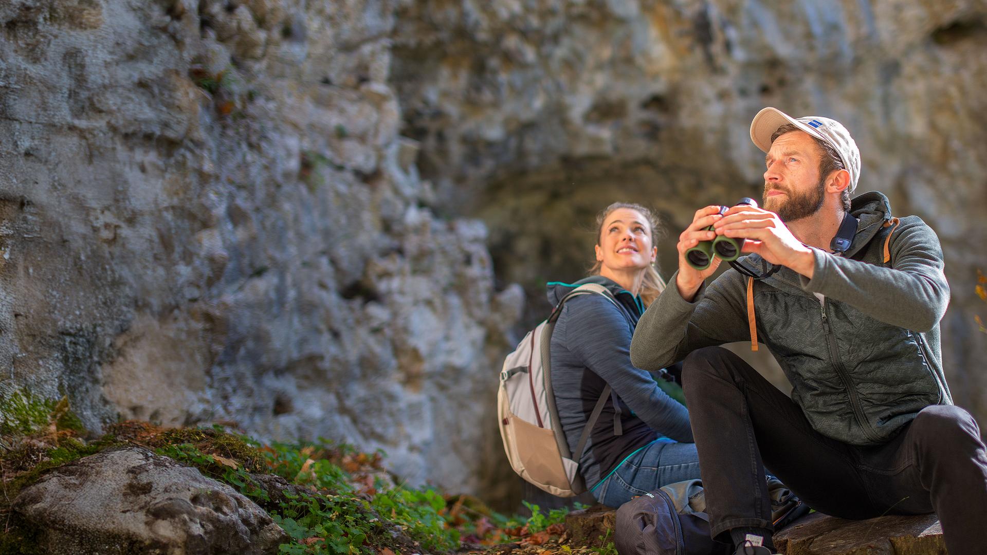 A man and woman in outdoor gear sit near a rocky cliff, with the man holding ZEISS binoculars and looking into the distance.