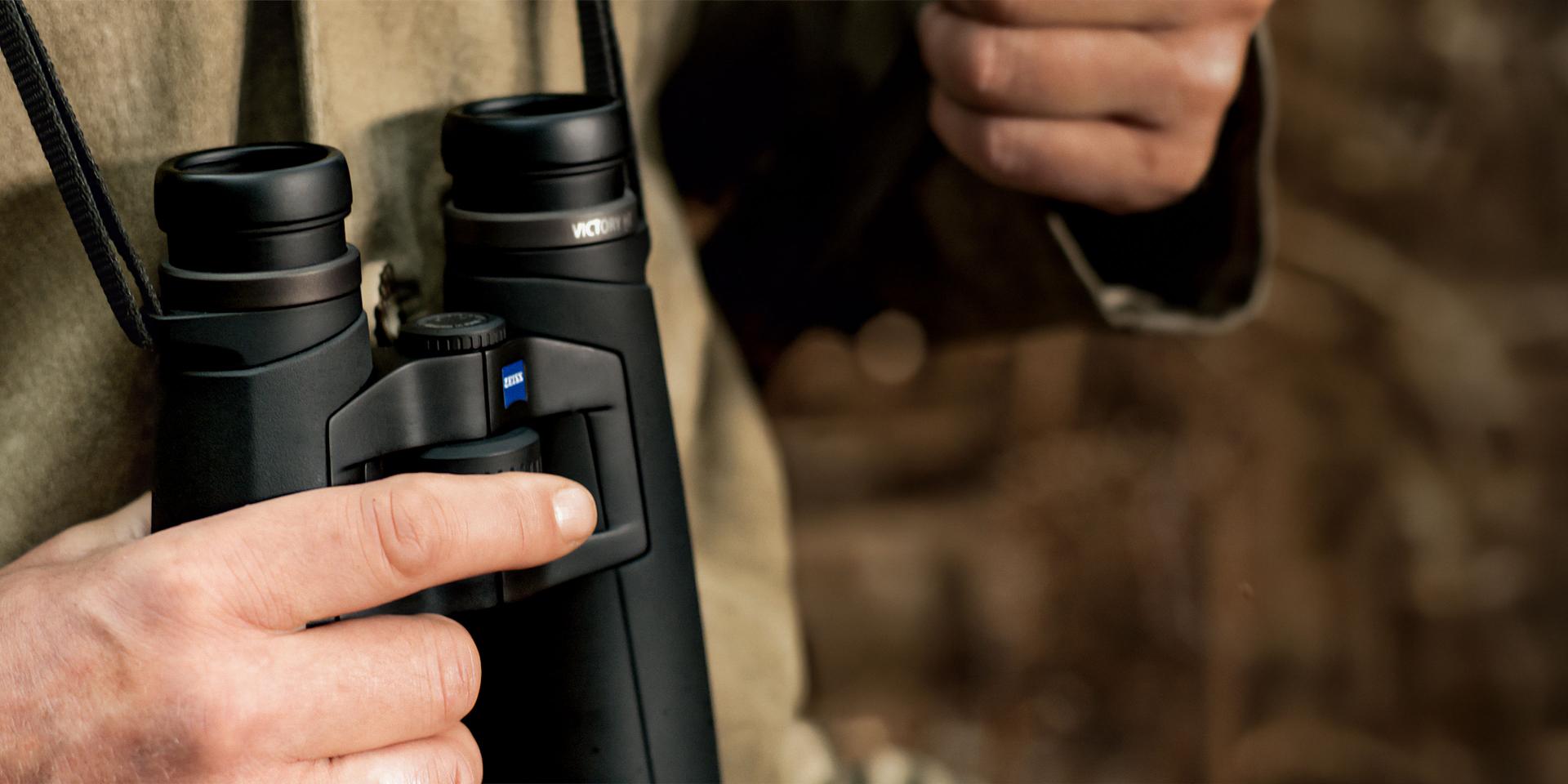 Person holding a pair of ZEISS binoculars with both hands outdoors.
