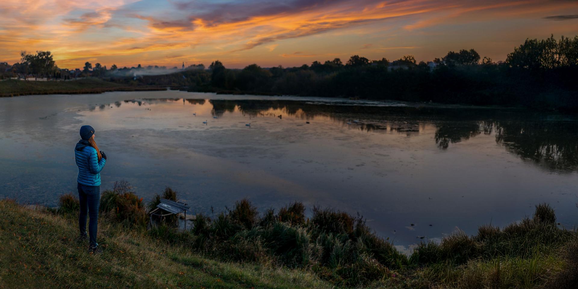 A person stands by a calm lake at sunrise using ZEISS binoculars to observe the natural landscape.