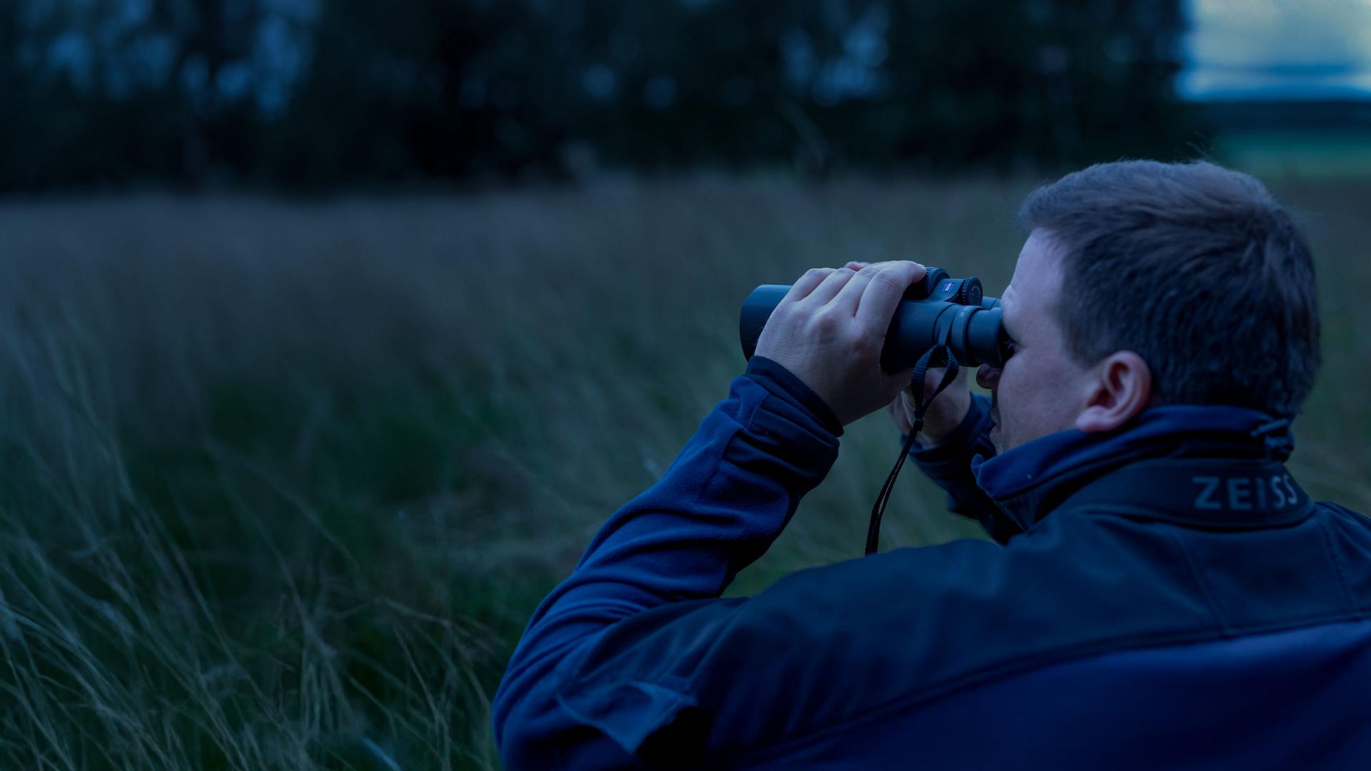 A person wearing a jacket is observing a field through ZEISS binoculars at dusk.