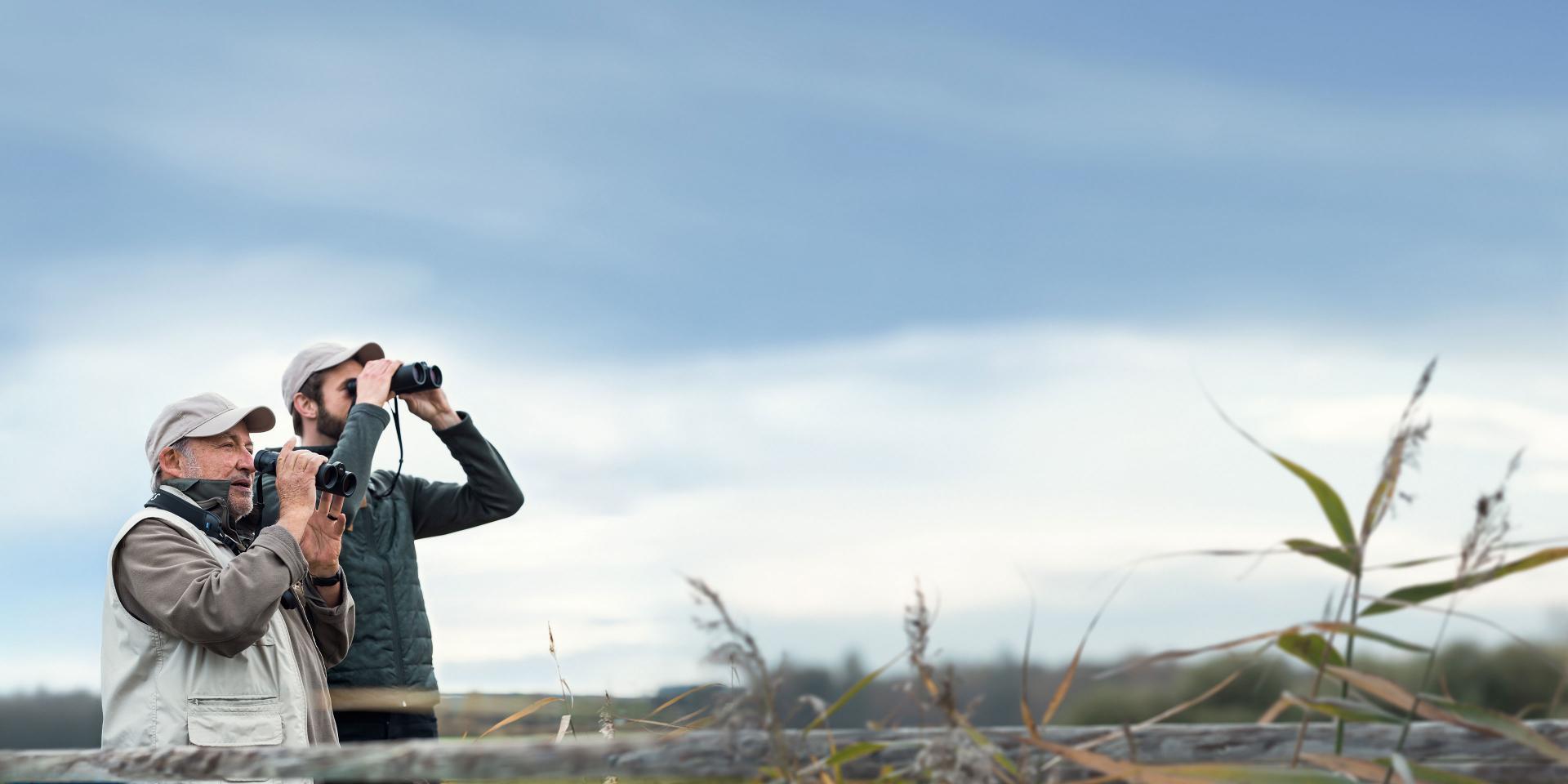 Two people are observing the landscape with ZEISS binoculars in a natural outdoor setting.