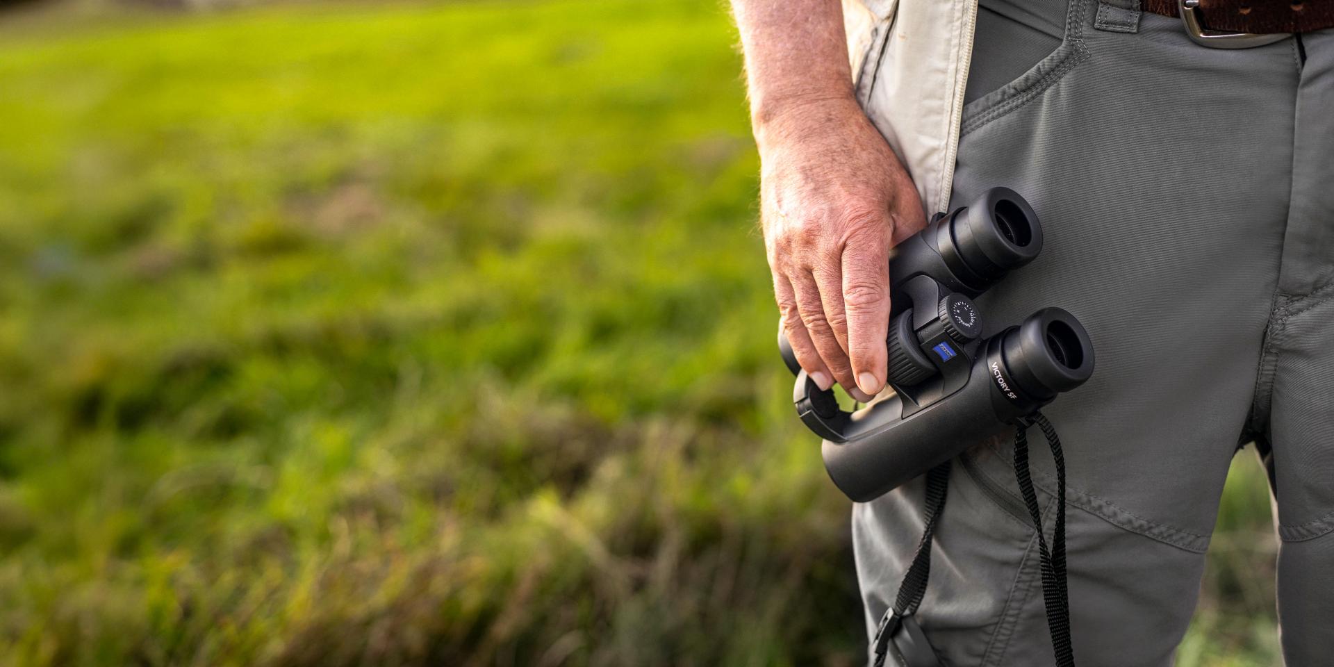 A person standing in a grassy field is holding a pair of ZEISS binoculars in one hand.