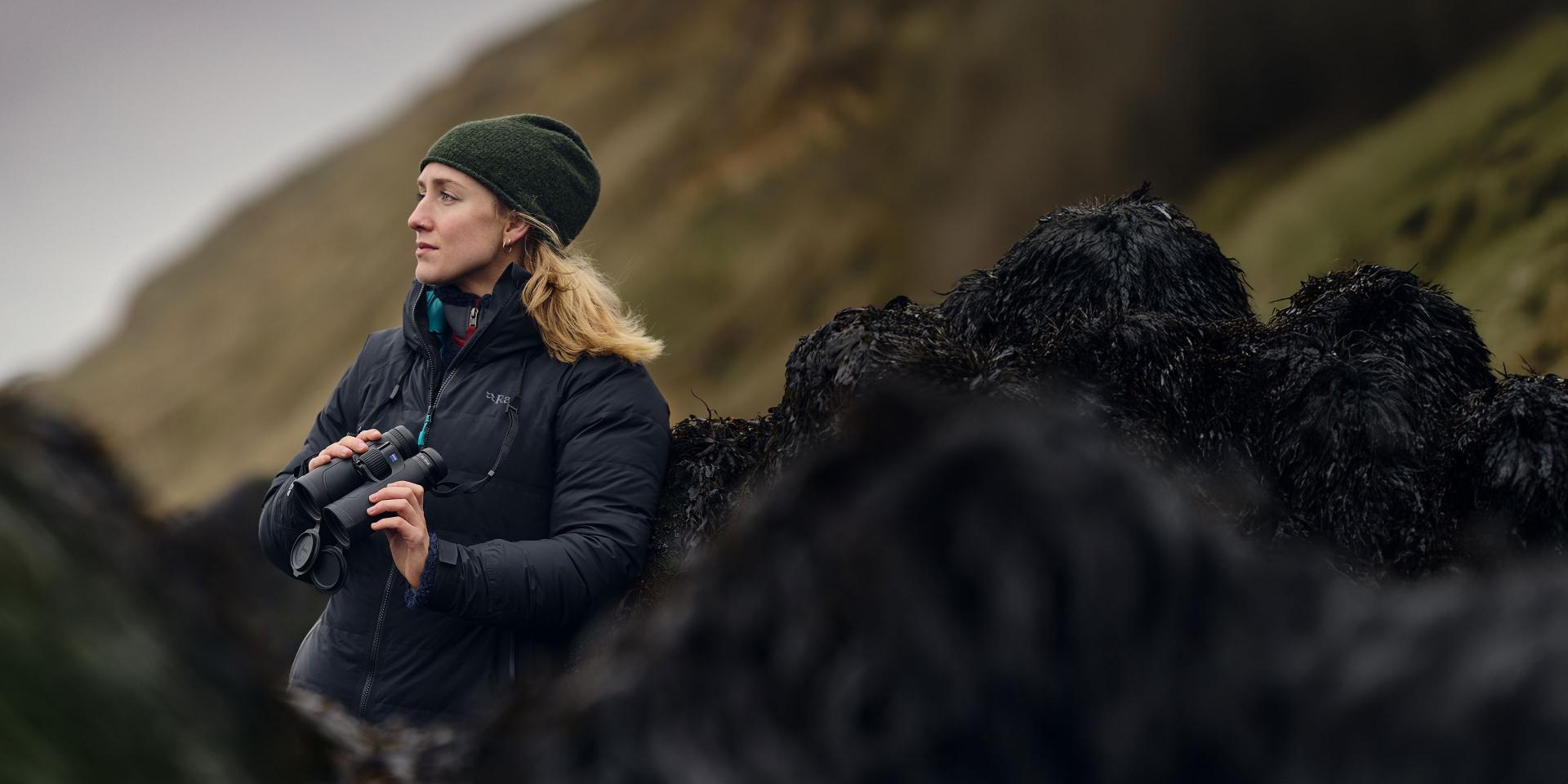 A woman in outdoor clothing stands on a rocky hillside holding a pair of ZEISS binoculars while looking into the distance.