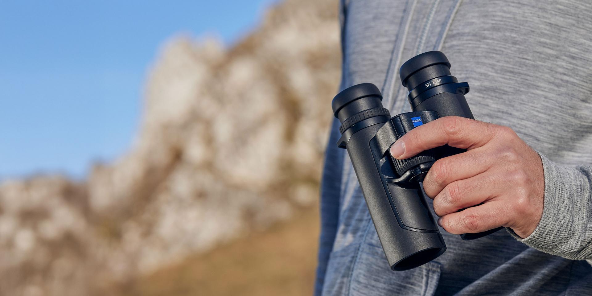 A person in a gray hoodie holds a pair of black ZEISS binoculars outdoors with a rocky mountain background.
