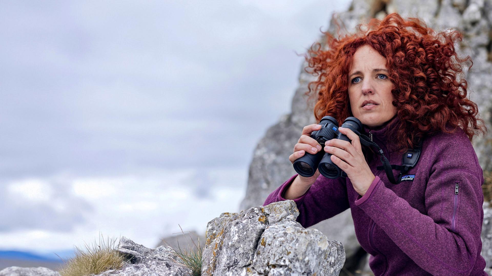 A woman with curly red hair uses ZEISS binoculars while observing the landscape from a rocky mountain.