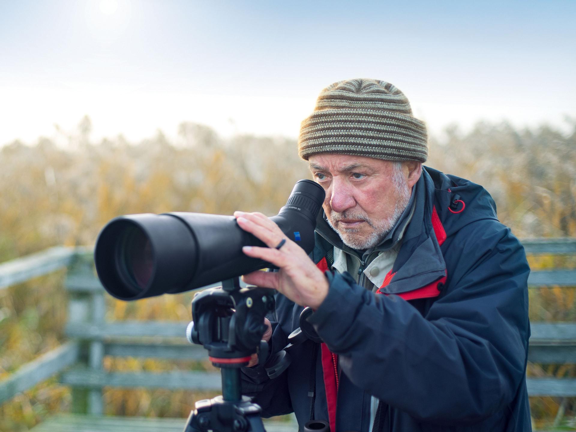 An older man using a ZEISS spotting scope mounted on a tripod for birdwatching in a natural outdoor setting.