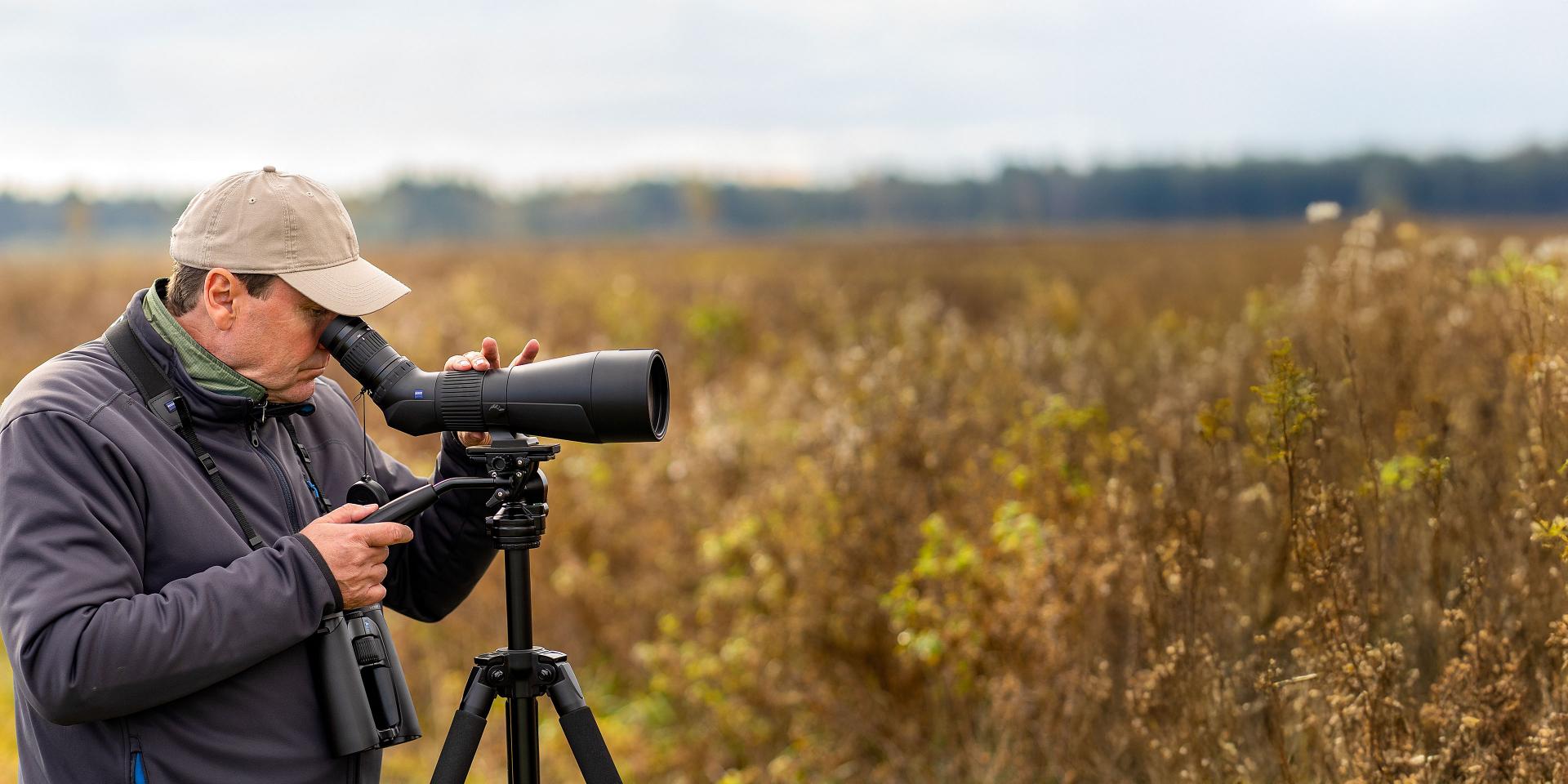 Man using a ZEISS spotting scope mounted on a tripod in a field with binoculars hanging from his neck.