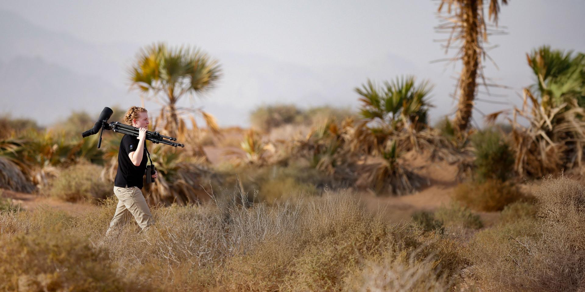 A person walks through a desert landscape carrying a tripod and ZEISS camera equipment on their shoulder.
