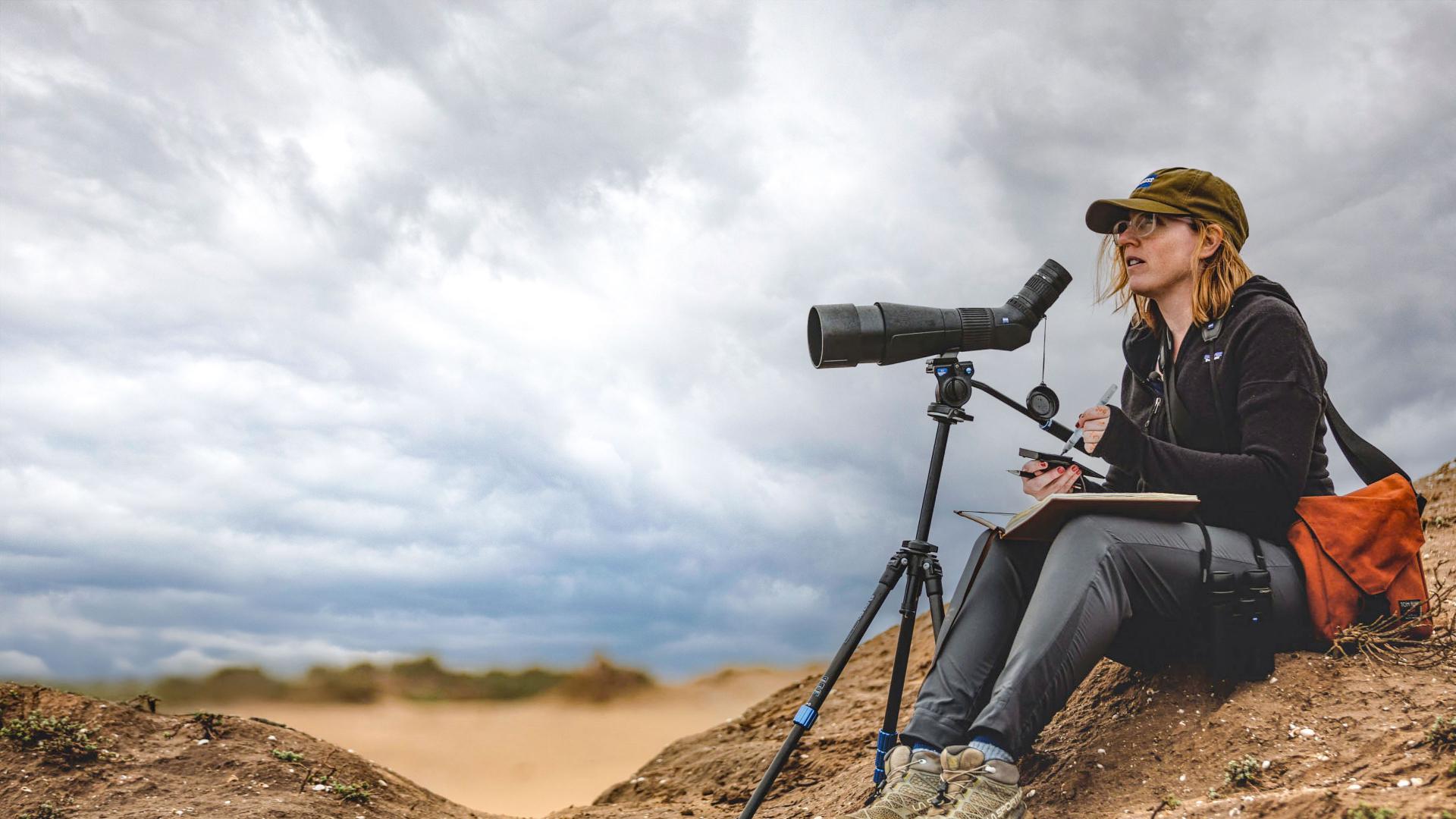 A woman sits outdoors in a sandy landscape using a ZEISS spotting scope mounted on a tripod while taking notes.