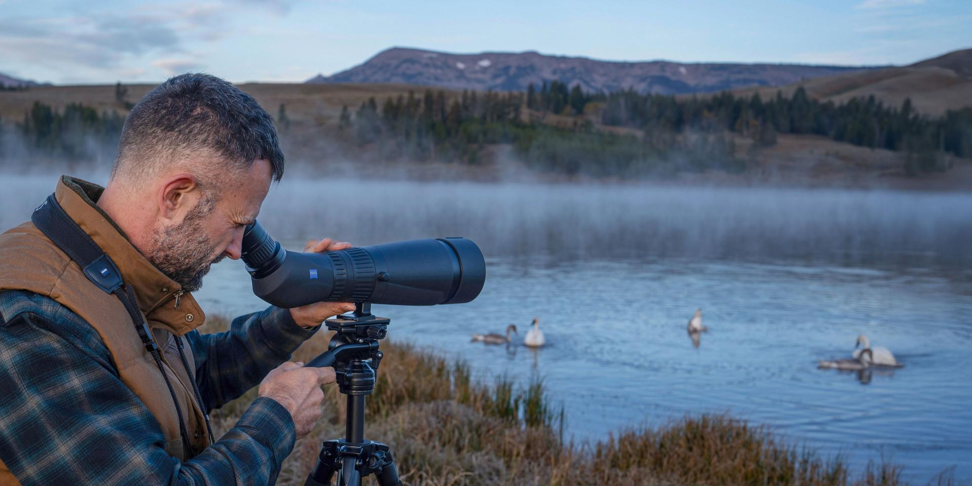 A man uses a ZEISS spotting scope on a tripod to observe swans on a misty lake in a mountainous landscape.