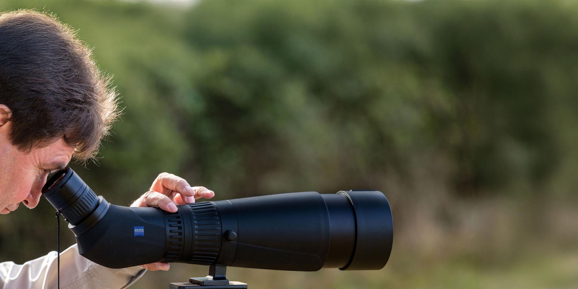 A person is looking through a ZEISS spotting scope mounted on a tripod in an outdoor setting.