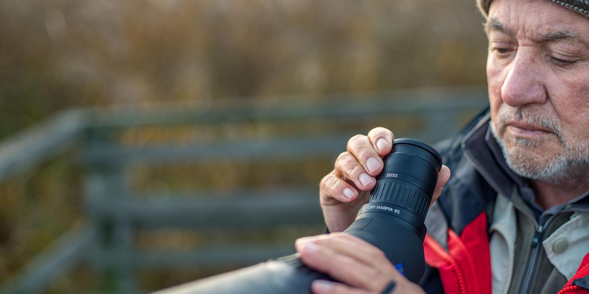 An older man in outdoor clothing adjusts the focus ring on a ZEISS spotting scope.