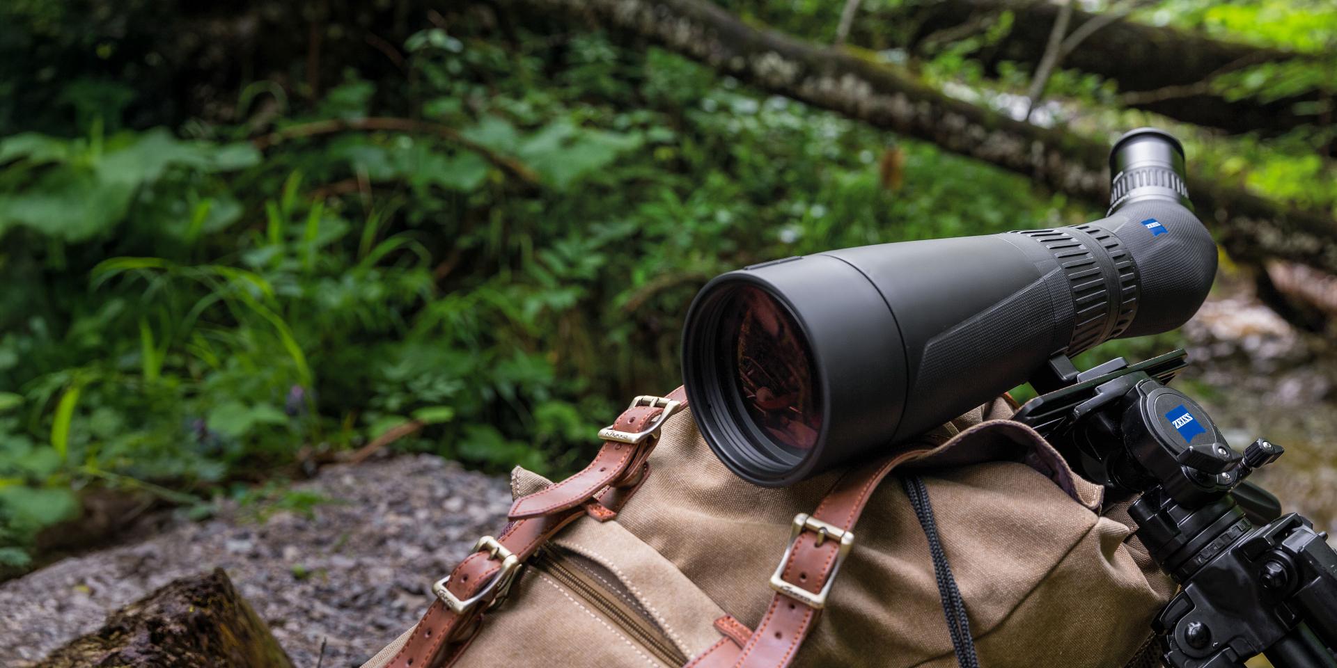 A ZEISS spotting scope mounted on a tripod rests on a backpack in a lush forest setting.