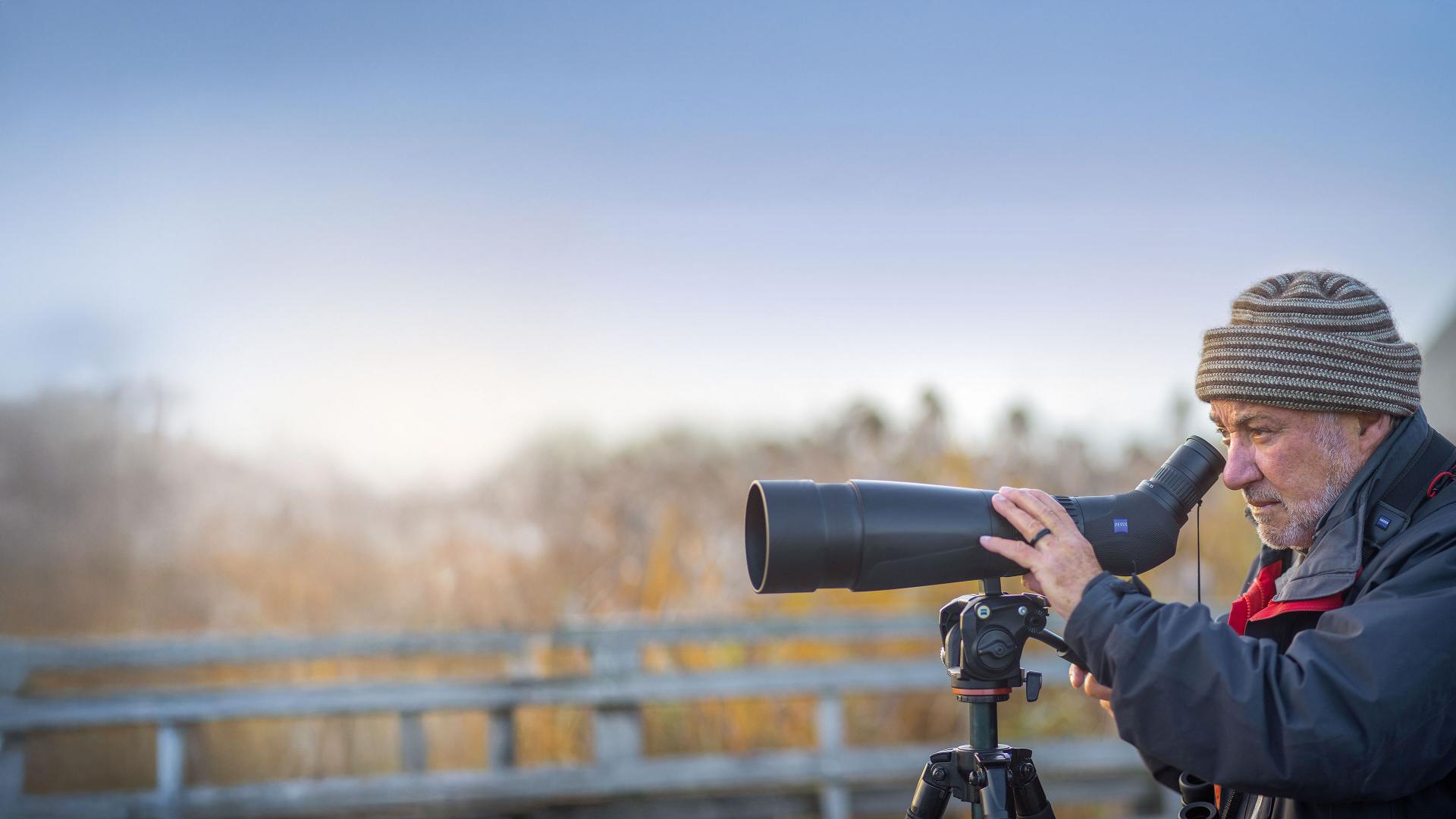 An older man observes nature through a ZEISS spotting scope mounted on a tripod in an outdoor setting.