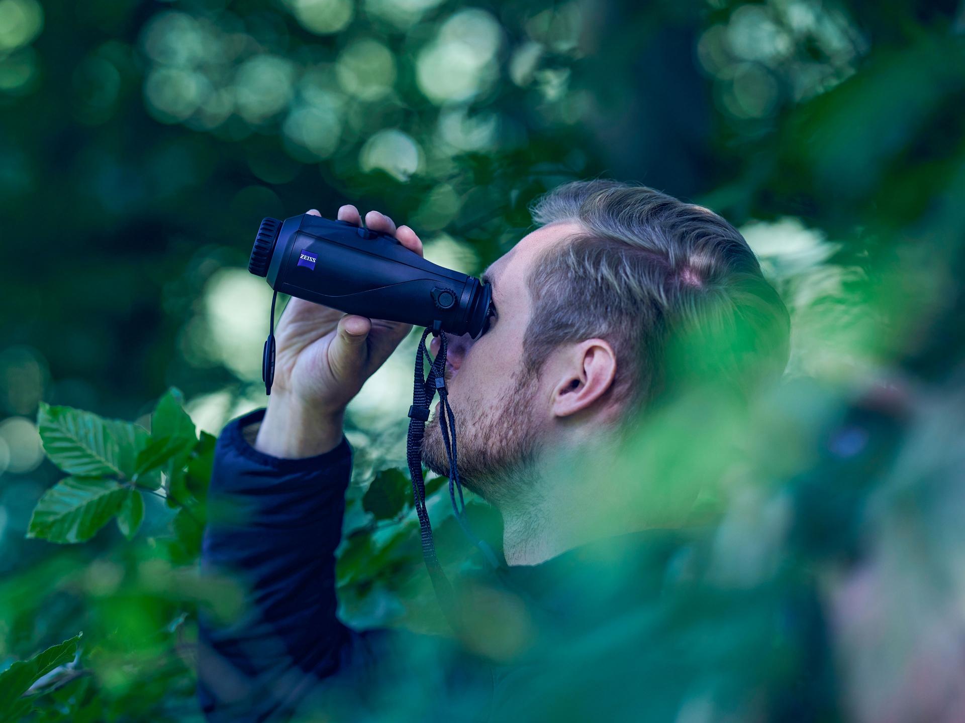 A man is observing through ZEISS binoculars in a lush, green forest environment.