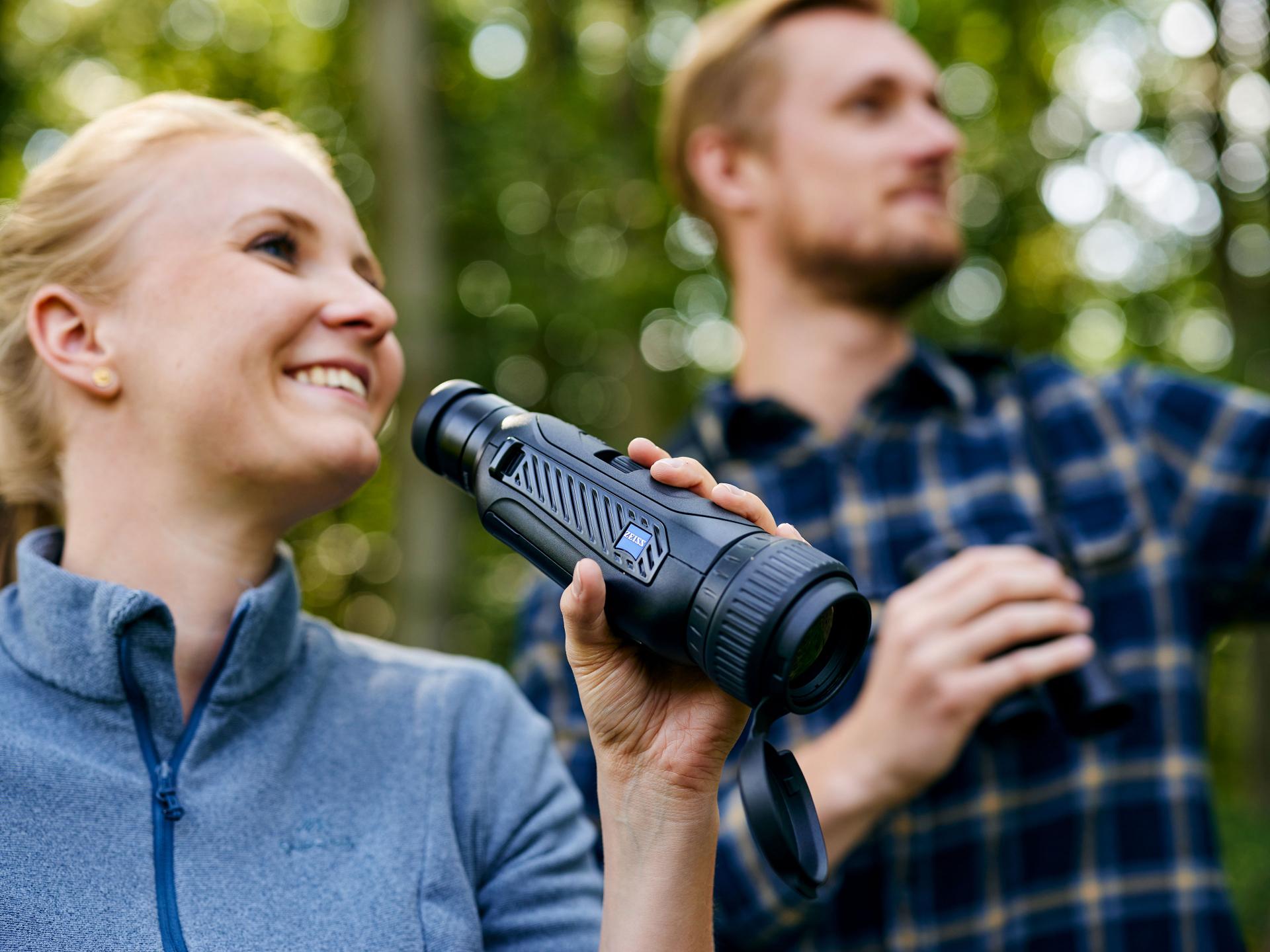 A woman holds a ZEISS thermal imaging device while a man stands beside her with binoculars in a forest setting.