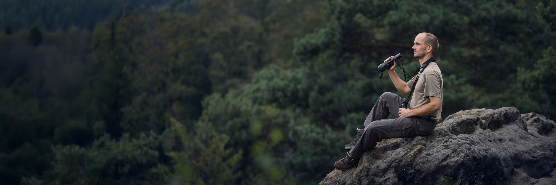 A man sitting on a rocky ledge in a forested area holding a pair of ZEISS binoculars.