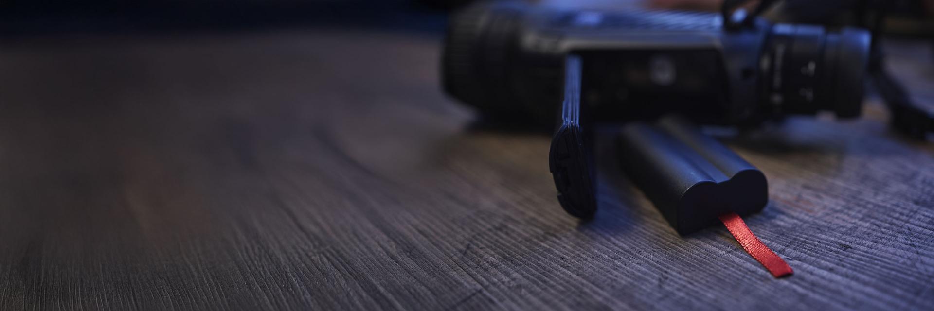 A ZEISS device lies on a wooden surface next to a removed battery with a red pull tab.