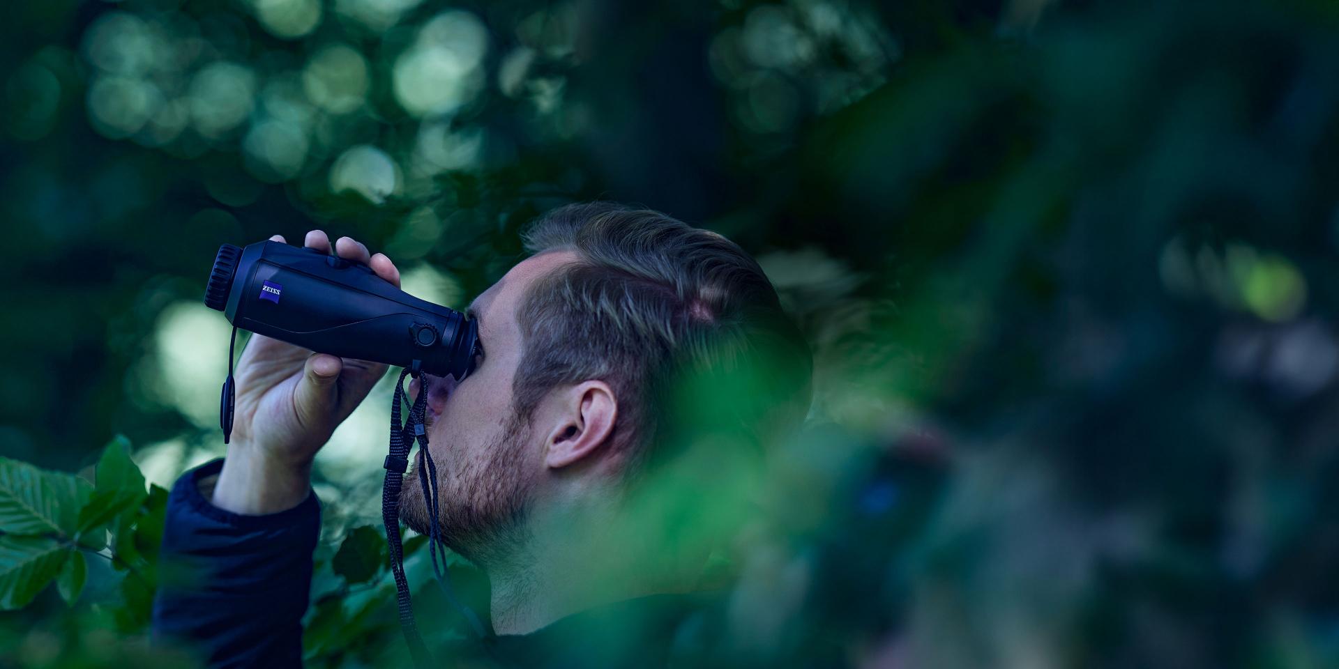 A man is observing through ZEISS binoculars in a forested area surrounded by green foliage.