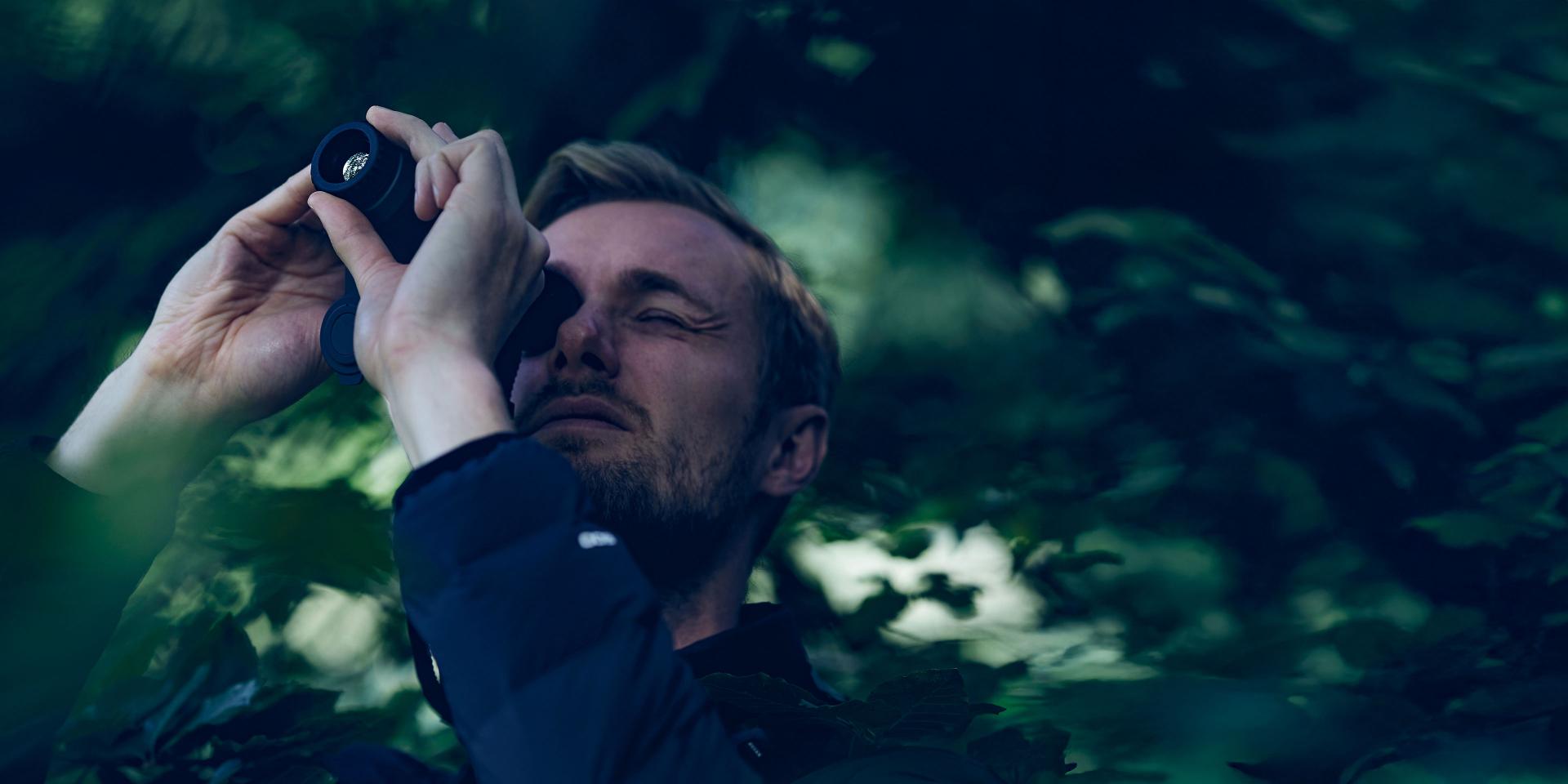 A man surrounded by dense foliage looks through a ZEISS monocular while observing nature.