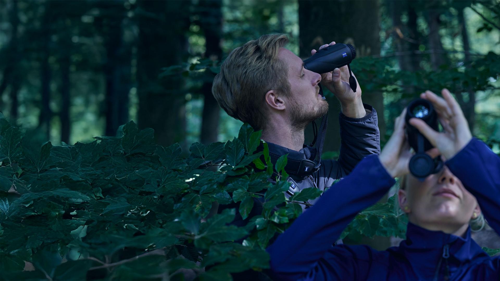 Two people are observing wildlife in a forest using ZEISS binoculars.