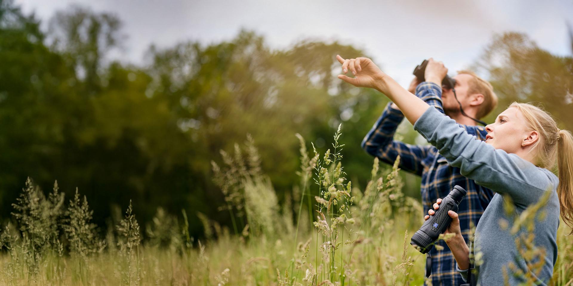 Two people standing in a grassy field using ZEISS binoculars to observe something in the sky.