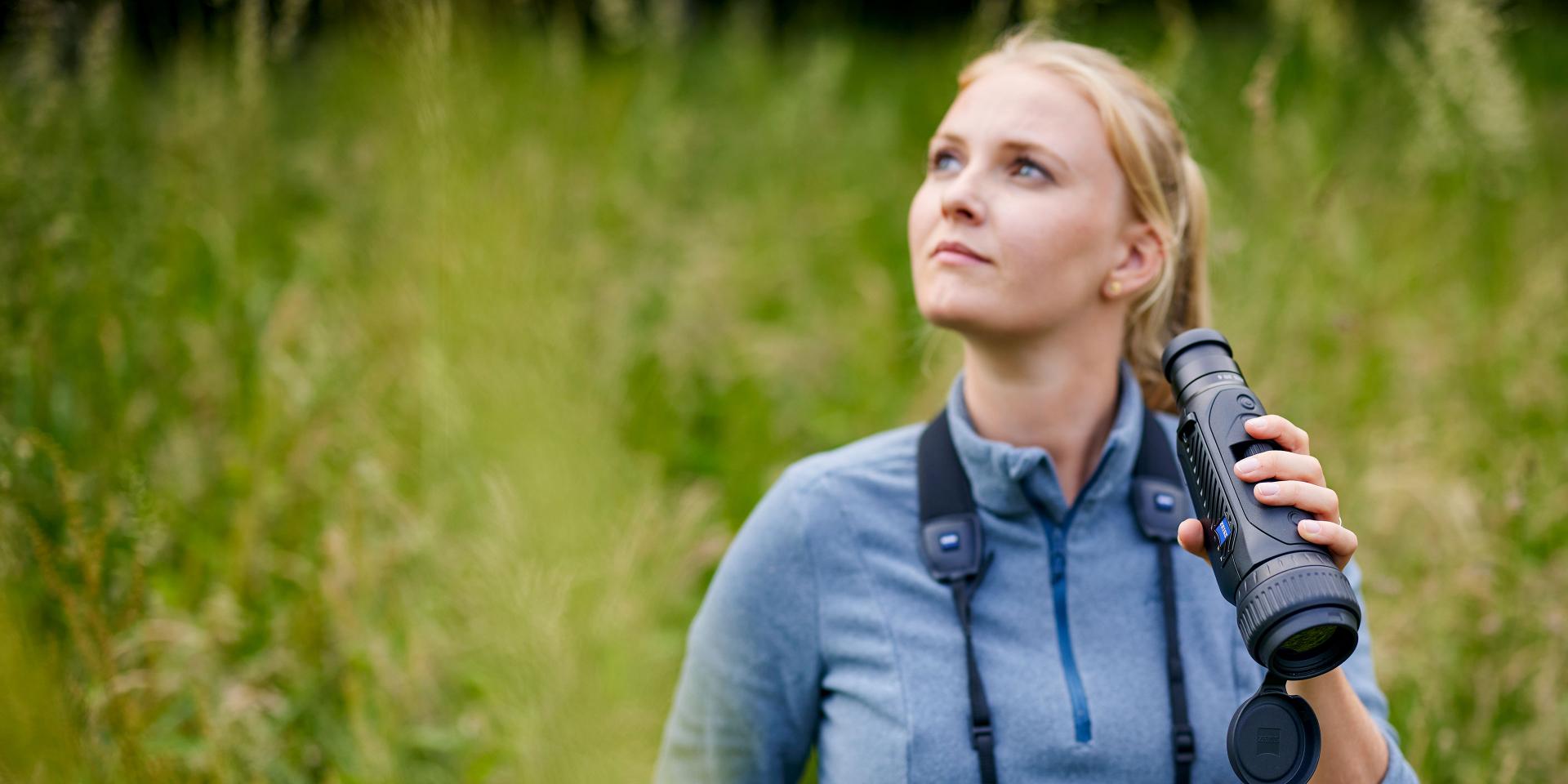 A woman standing in a grassy field holds a ZEISS spotting scope and looks upwards.