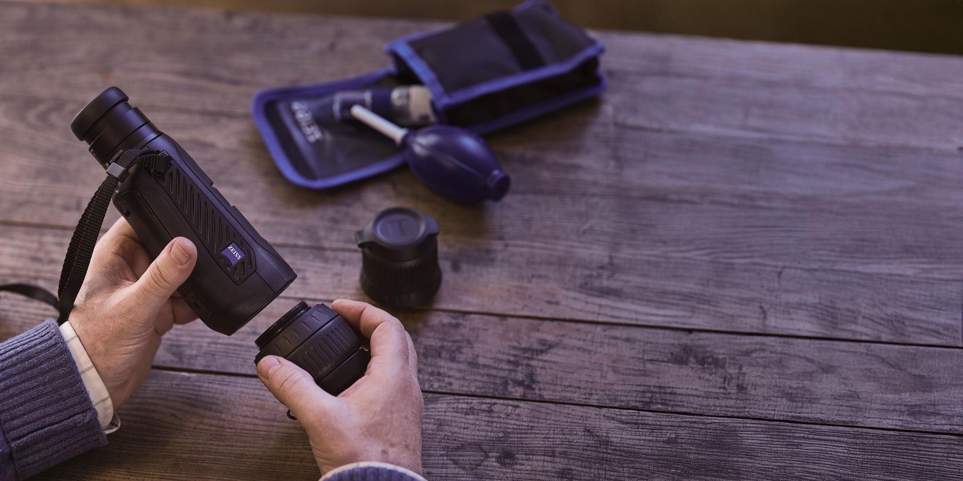 A person is assembling or cleaning a ZEISS monocular device on a wooden table with a ZEISS cleaning kit nearby.