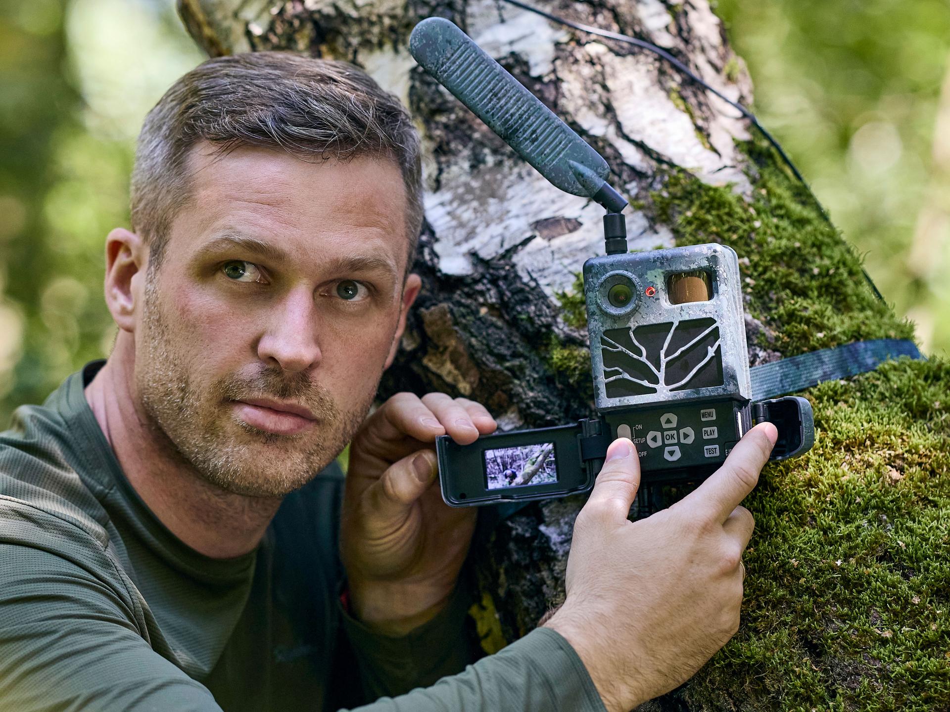 Man in outdoor clothing setting up a wildlife monitoring camera with a ZEISS device in a forest environment.