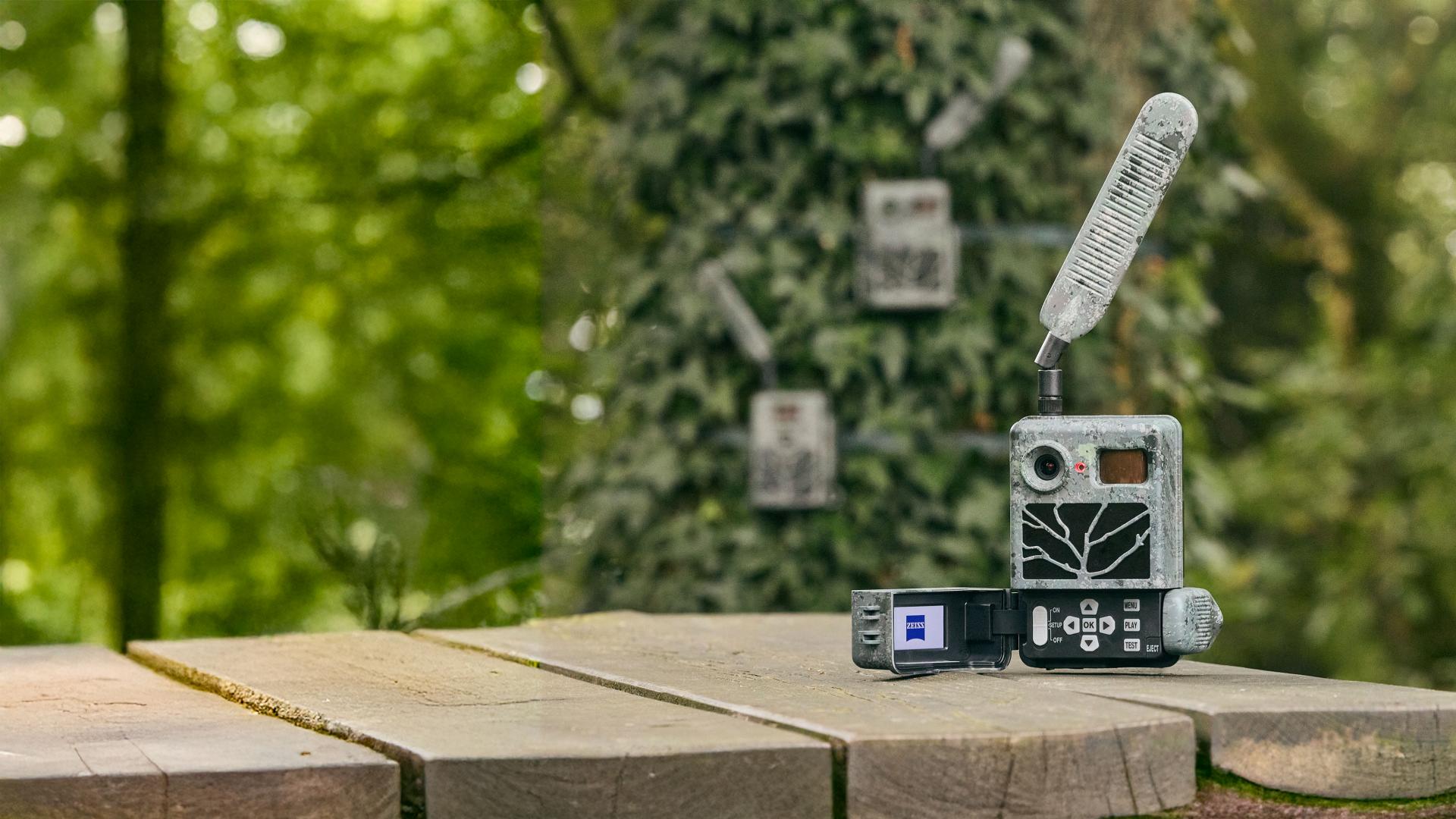A ZEISS trail camera and digital device are placed on a wooden surface in a forest setting with additional trail cameras mounted on a tree in the background.