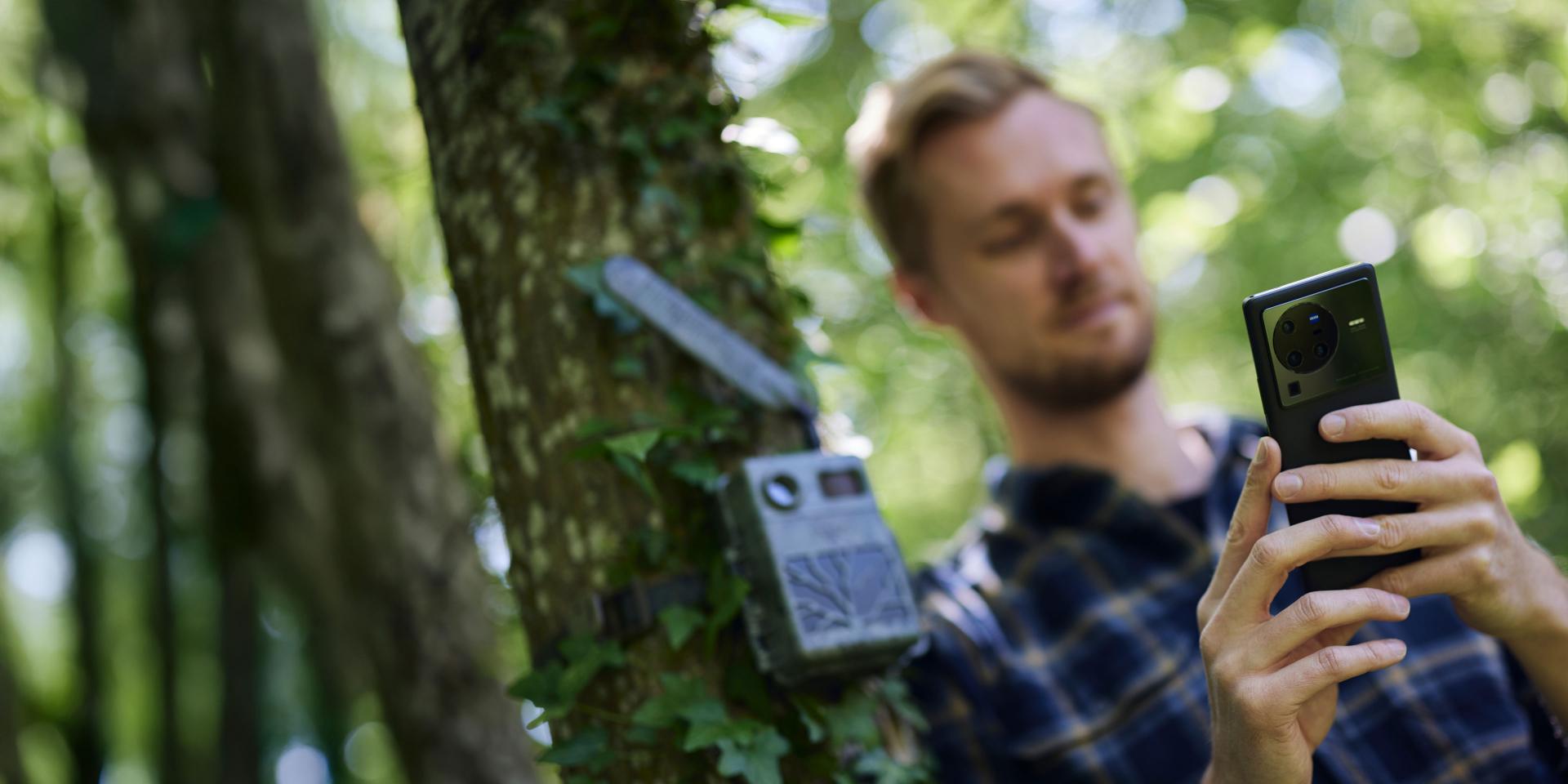 A man in a forest uses a smartphone with a ZEISS lens to photograph or scan a wildlife camera attached to a tree.