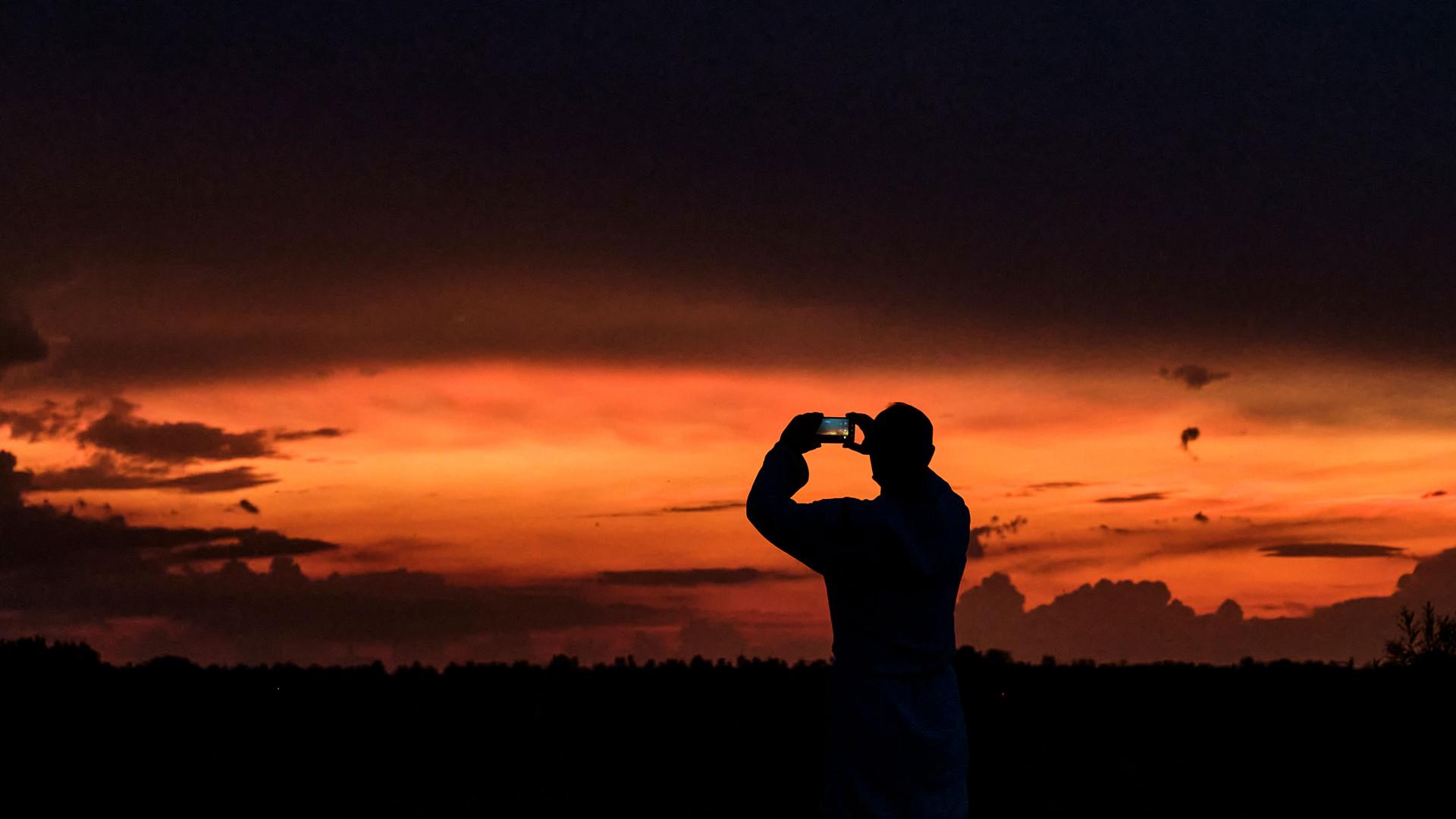 A person silhouetted against a vivid orange sunset sky takes a photo using a smartphone equipped with a ZEISS lens.