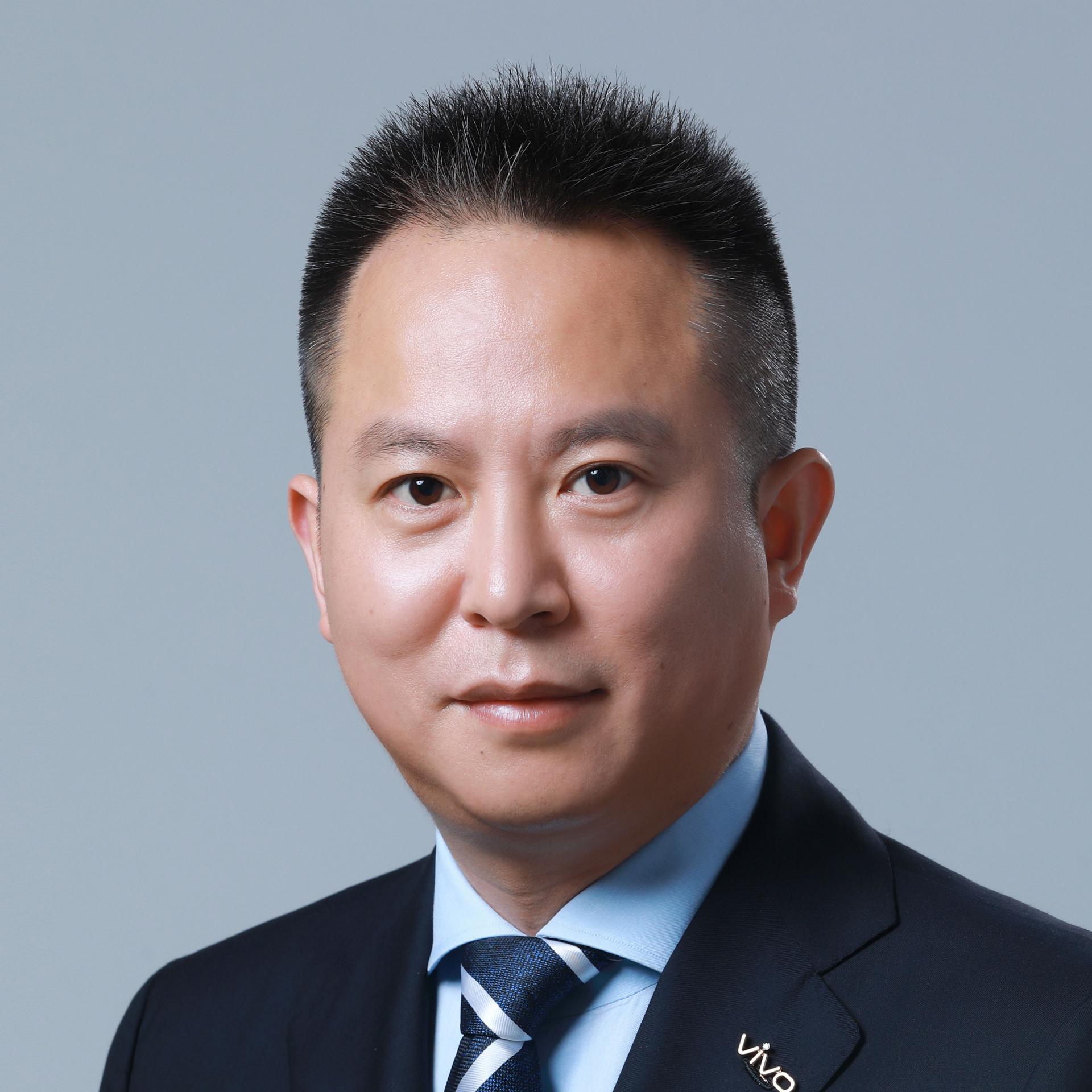 Portrait of a man in a dark suit and striped tie, wearing a lapel pin with the "vivo" logo, against a plain light grey background.