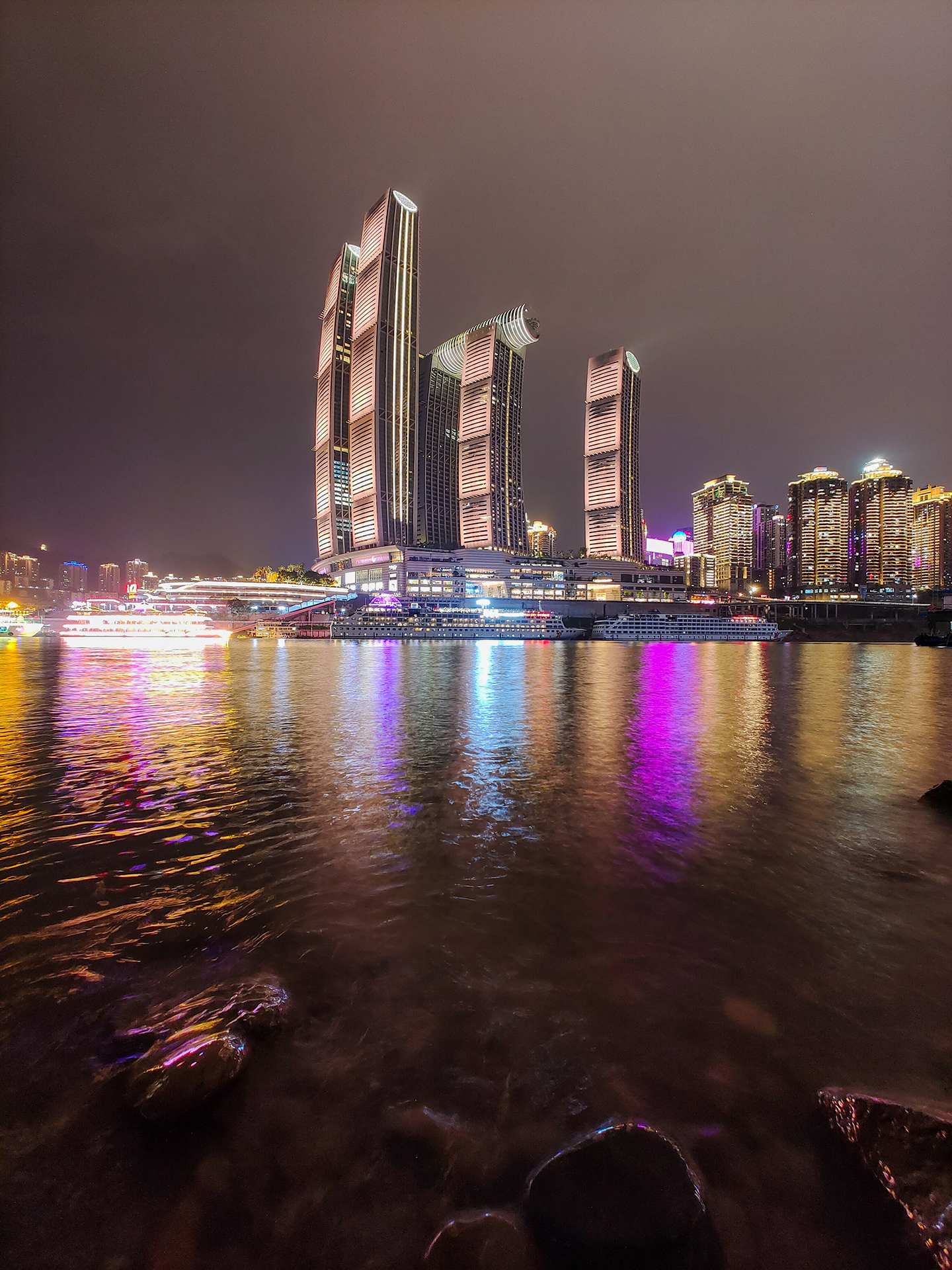 Modern high-rise buildings illuminated at night are reflected in the calm water of a river, showcasing vibrant city lights and architectural detail captured with ZEISS clarity.