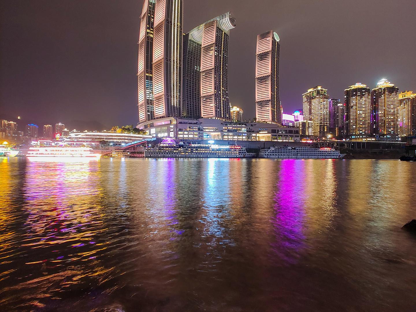 Modern high-rise buildings illuminated at night are reflected in the calm water of a river, showcasing vibrant city lights and architectural detail captured with ZEISS clarity.