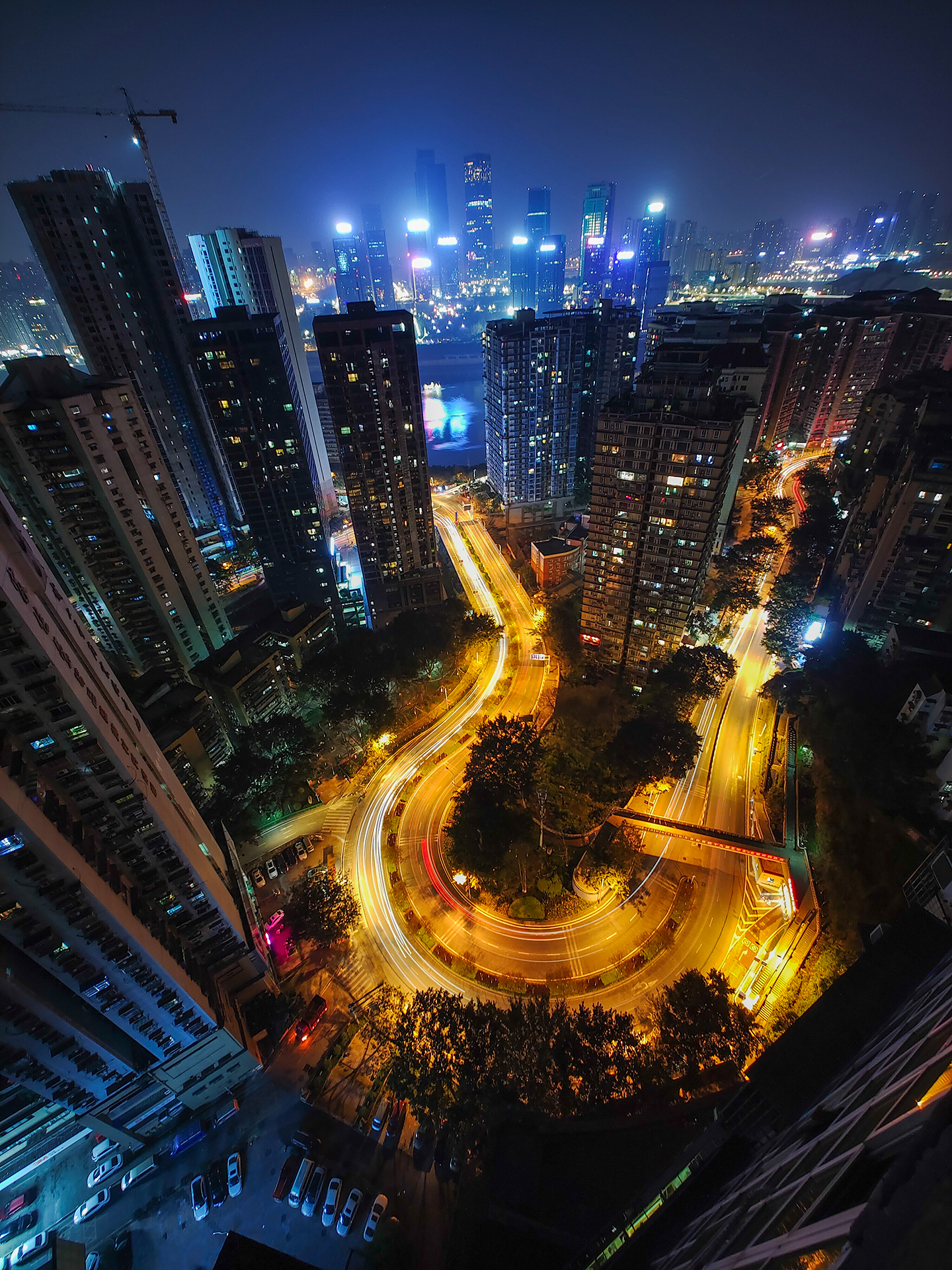 Nighttime cityscape captured from a high vantage point, showcasing tall buildings, winding illuminated roads, and a glowing skyline in the distance, demonstrating ZEISS lens clarity and dynamic low-light performance.