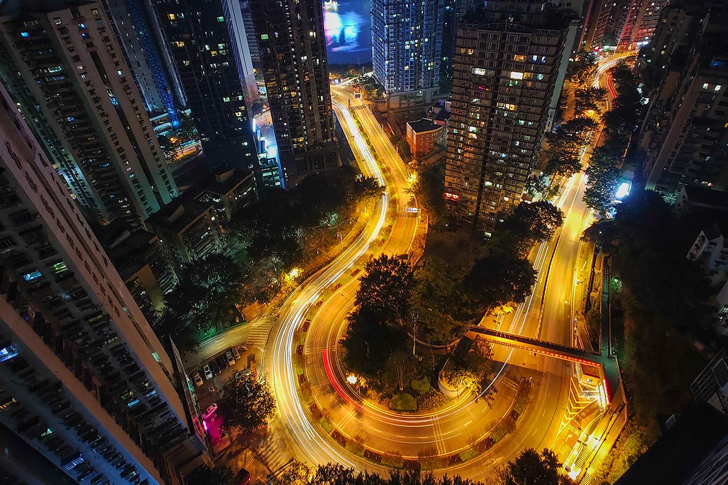 Nighttime cityscape captured from a high vantage point, showcasing tall buildings, winding illuminated roads, and a glowing skyline in the distance, demonstrating ZEISS lens clarity and dynamic low-light performance.