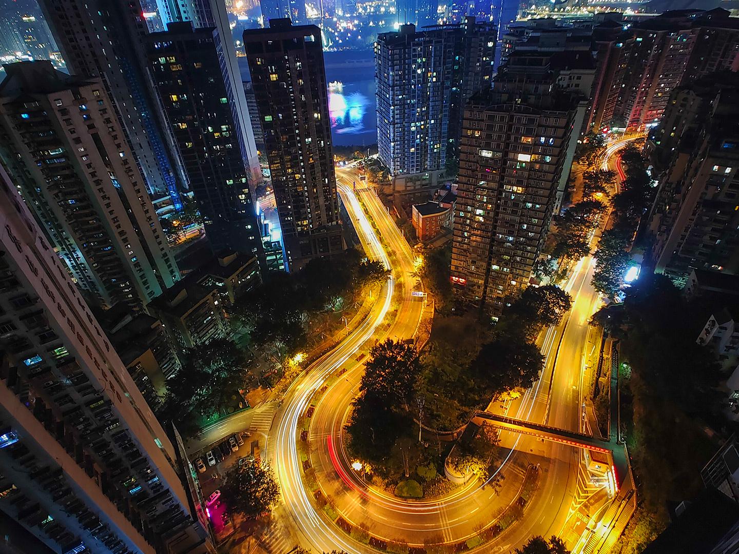 Nighttime cityscape captured from a high vantage point, showcasing tall buildings, winding illuminated roads, and a glowing skyline in the distance, demonstrating ZEISS lens clarity and dynamic low-light performance.