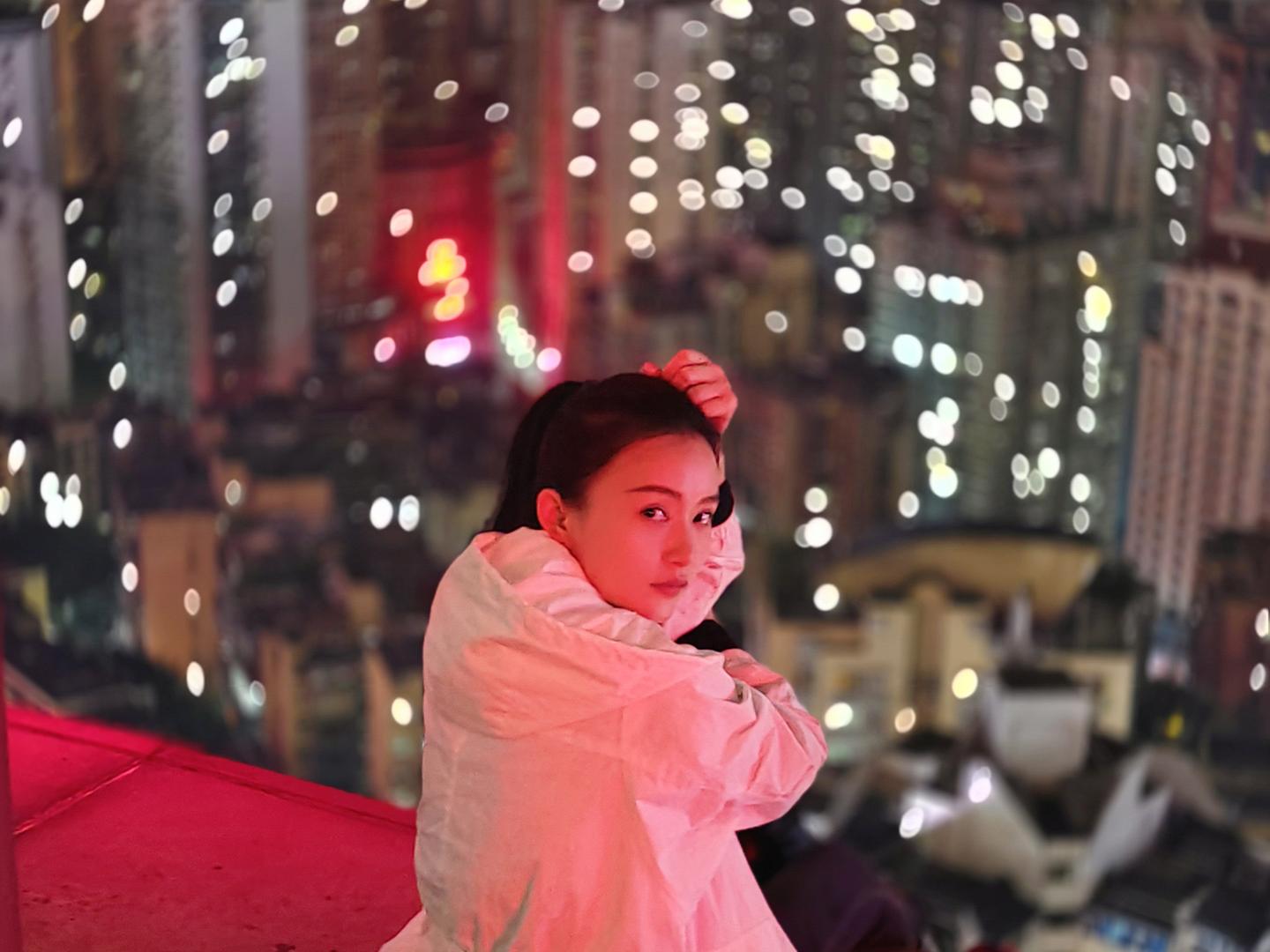 A woman in a white coat sits on a rooftop ledge at night with a vibrant, bokeh-filled cityscape in the background, showcasing ZEISS lens clarity and depth.