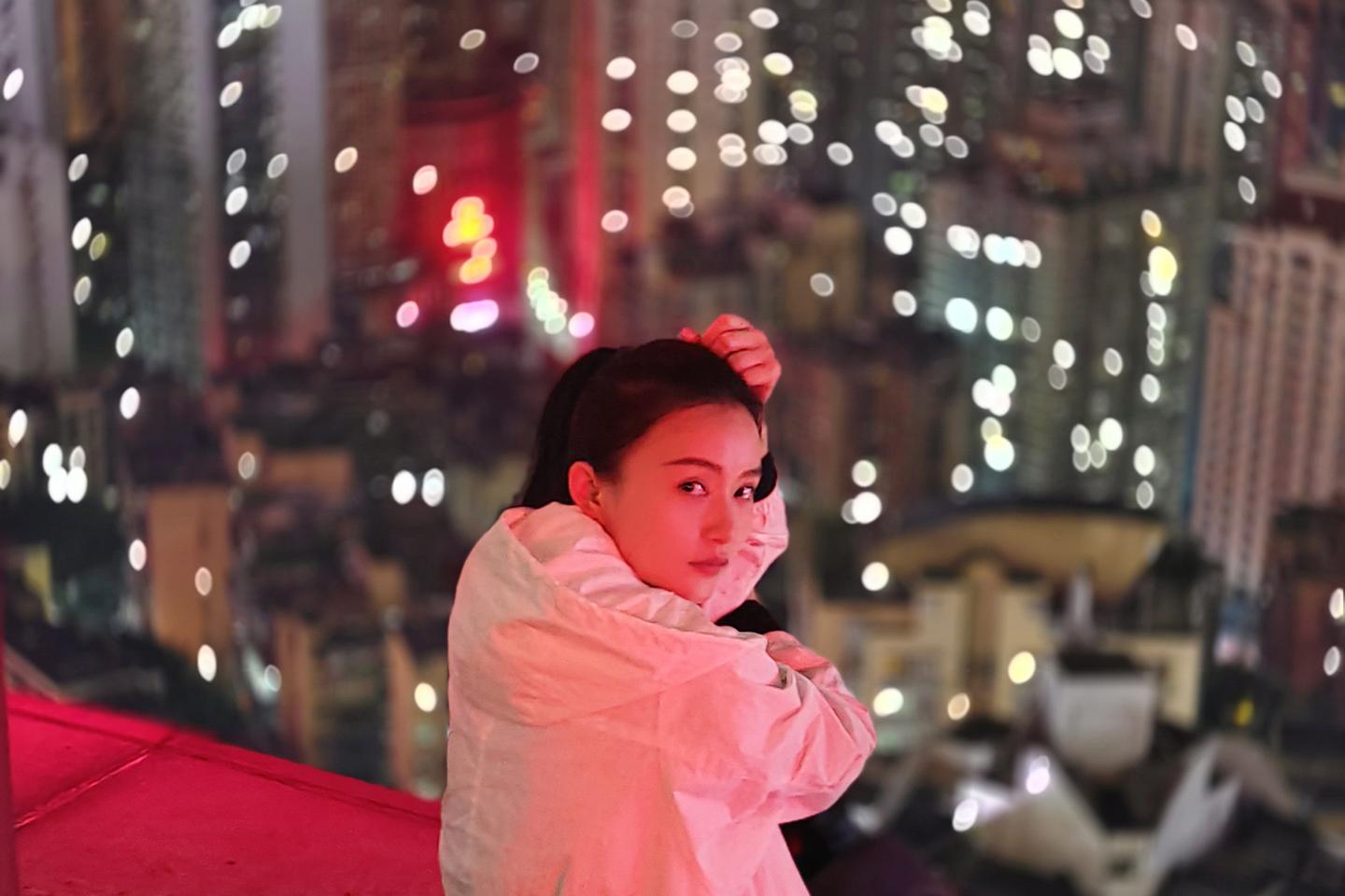 A woman in a white coat sits on a rooftop ledge at night with a vibrant, bokeh-filled cityscape in the background, showcasing ZEISS lens clarity and depth.
