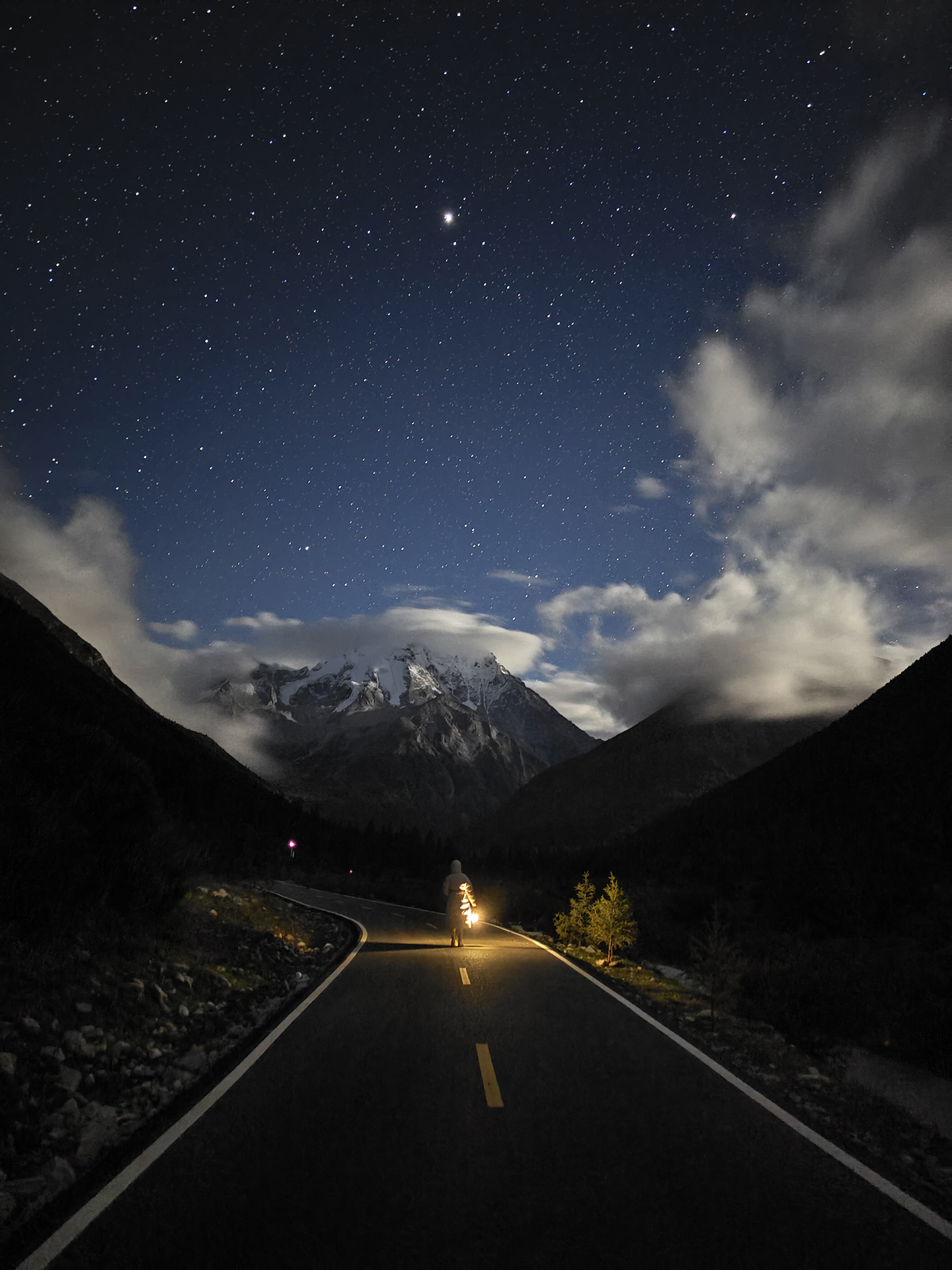 A lone man stands on a winding mountain road under a star-filled night sky with snow-capped peaks in the background, captured with stunning clarity and contrast.