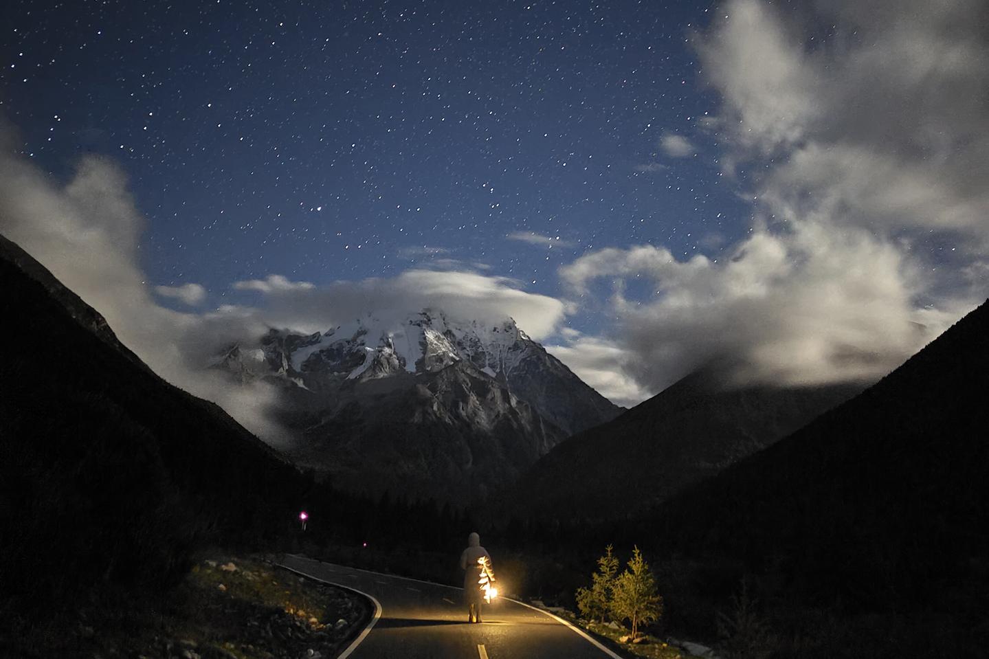 A lone man stands on a winding mountain road under a star-filled night sky with snow-capped peaks in the background, captured with stunning clarity and contrast.